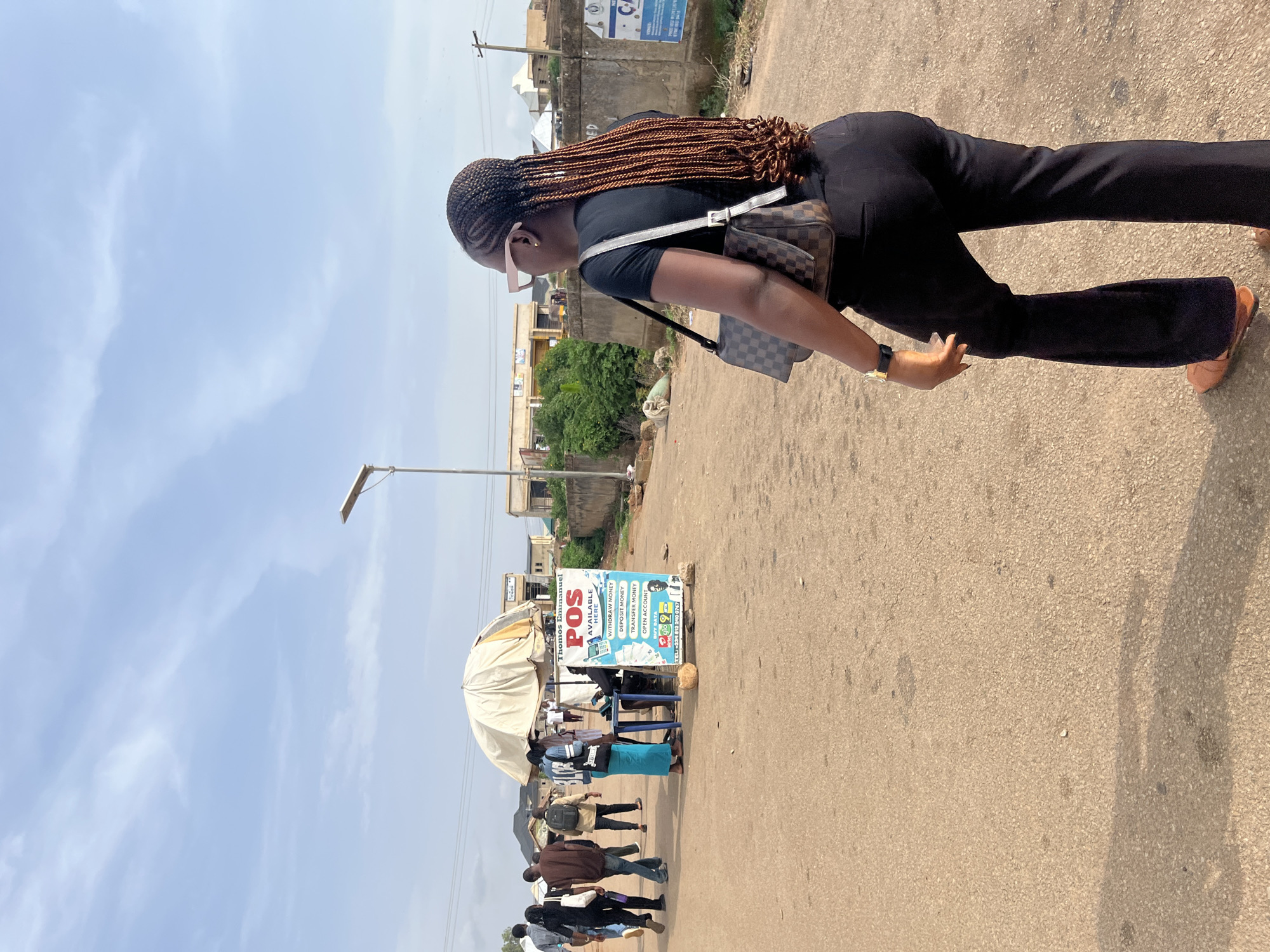In an outdoor urban setting, possibly in Rafin Par, Nigeria, a woman with braided hair, wearing a black t-shirt, black flared pants, and brown sandals, walks away from the camera. She carries a brown and checkered handbag across her body and a gold wristwatch on her left wrist. In the background, several people are seen walking on a paved road. To the right of the road, there is a stall with a sign advertising "Thomos Emmanuel POS AVAILABLE HERE" offering services like withdrawal, deposit, transfer, and account opening. A man sits behind the table at the stall. The environment appears to be an open area with a few buildings and vegetation visible in the distance, under a partly cloudy sky. A lamppost stands on the side of the road.