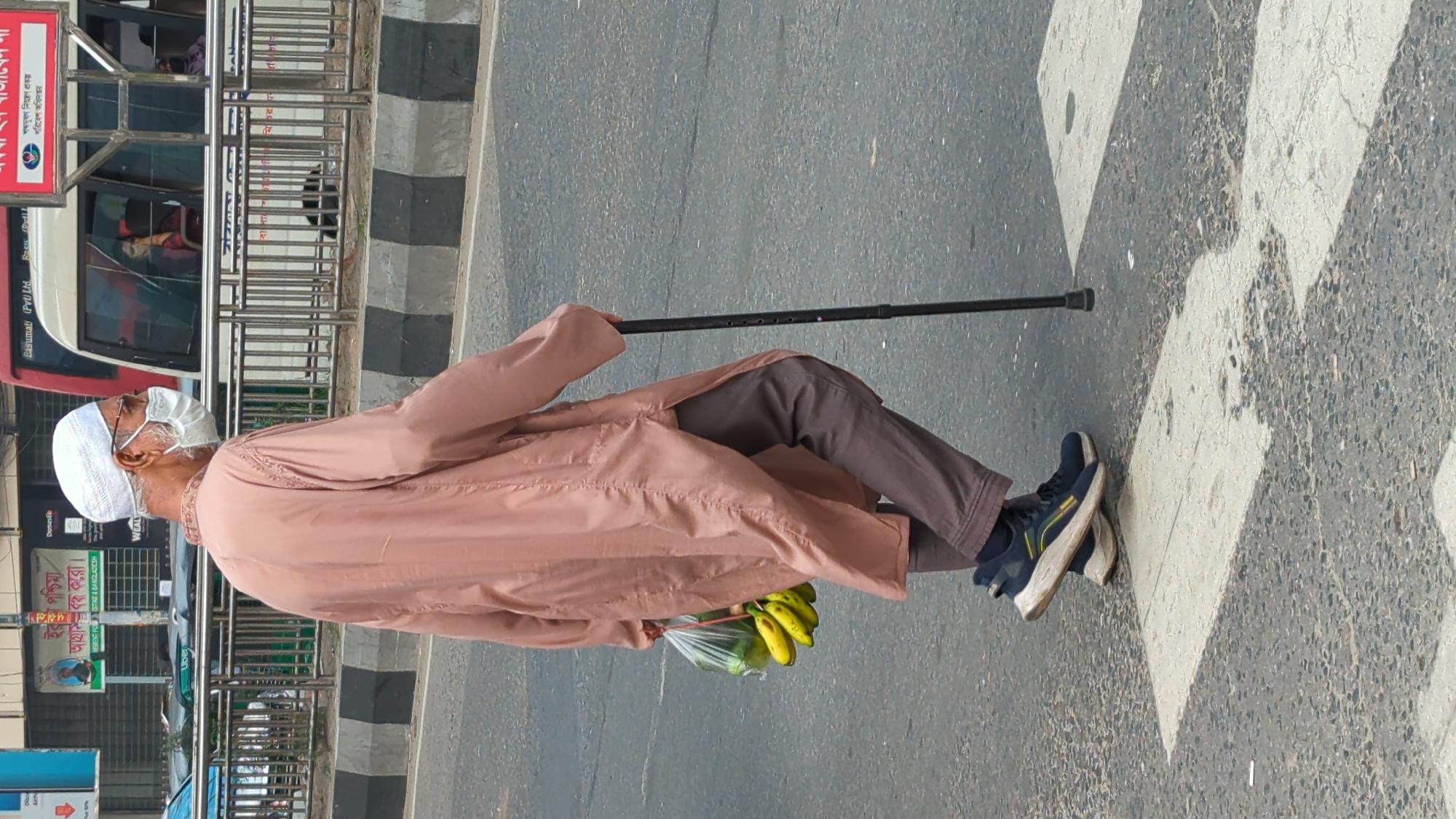 An elderly man is walking on a zebra crossing in Dhaka, Bangladesh. He is wearing a white cap, a face mask, a light brown robe, and brown trousers. He is carrying a plastic bag containing bananas in one hand and using a black walking stick with the other. The road surface is grey asphalt with white stripes. A bus with advertisements can be seen in the background.