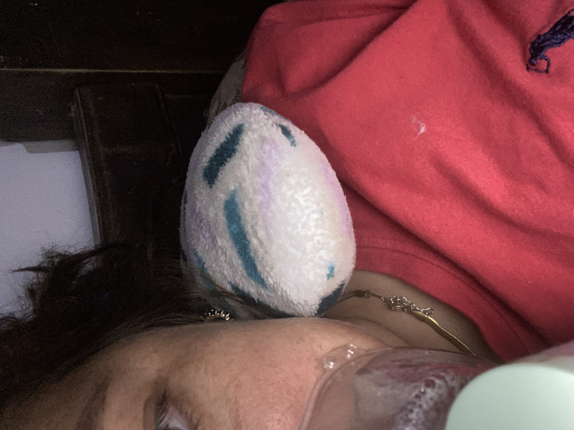 The image shows a close-up of a person lying down, with their head resting on a dark surface. They are wearing a red shirt with a navy blue embroidered logo. A fuzzy, egg-shaped object with blue and purple stripes is positioned near their head. A delicate gold necklace is visible around their neck, partially obscured by the red shirt. The lighting suggests an indoor setting, possibly at night or in a dimly lit room. The person's face is partially visible, with their eyes closed. The overall mood is serene and relaxed. The focus is on the textures of the objects and the person's relaxed state.