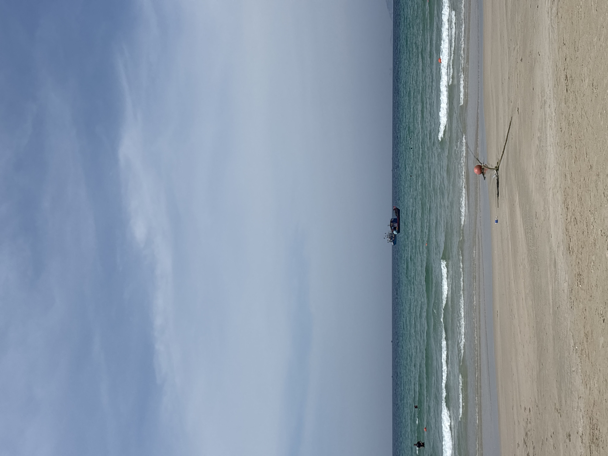The image depicts a serene beach scene with a sandy shore in the foreground. The ocean, with its turquoise waters and white-capped waves, stretches towards the horizon. Two fishing boats are visible in the distance, navigating the calm sea under a partly cloudy sky. A red buoy is anchored to the shore with a rope, signifying a boundary or marker. In the shallow water, a few individuals can be seen enjoying the beach. The overall atmosphere is peaceful and tranquil.