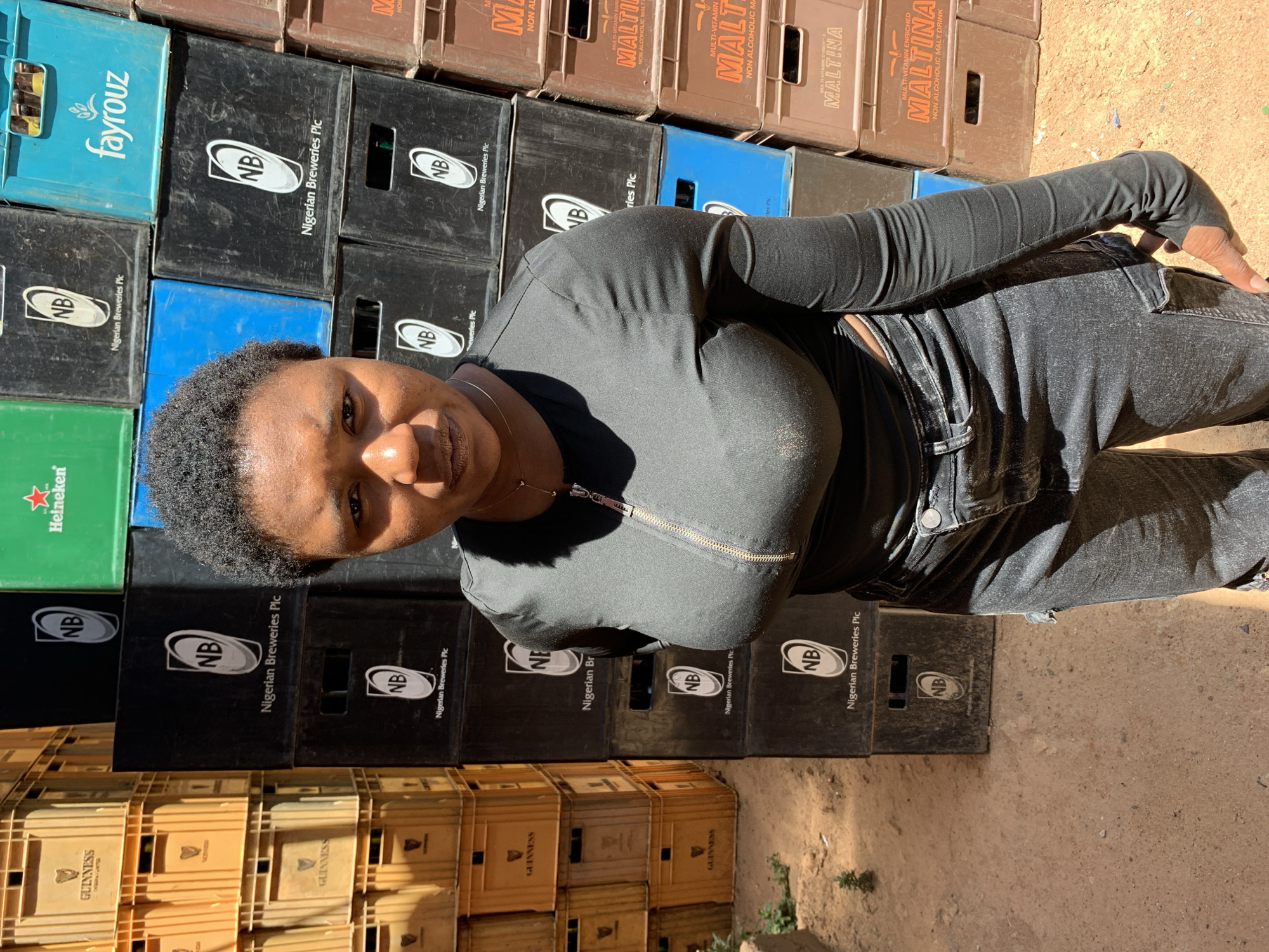 The image presents a woman posing in Gura Top, Nigeria, in front of stacked crates. The crates feature brands like Guinness, Nigerian Breweries Plc (NB), Fayrouz, Heineken, and Maltina. The woman is wearing a black long-sleeved top and dark jeans. The setting appears to be outdoors with a dirt ground.
