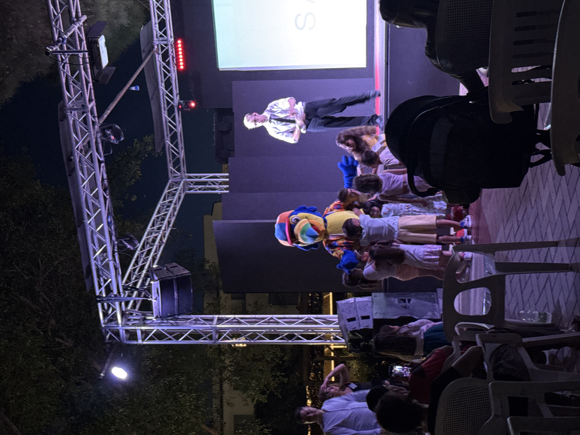 In Isola delle Femmine, Italy, a nighttime outdoor event is taking place on a stage with a metallic truss structure overhead. A person in a colorful parrot mascot costume is interacting with a group of children on stage. To the right of the mascot, a man in a white collared shirt and dark pants, possibly an entertainer or host, stands with his arms crossed. Behind them, a large screen displays a bright, illuminated area, partially obscured by a dark structure. In the foreground, an audience watches the performance from chairs, with a baby stroller visible. The ground is paved with brick-like tiles, and trees are present in the background. The scene is illuminated by stage lights and ambient lighting, creating a festive atmosphere.