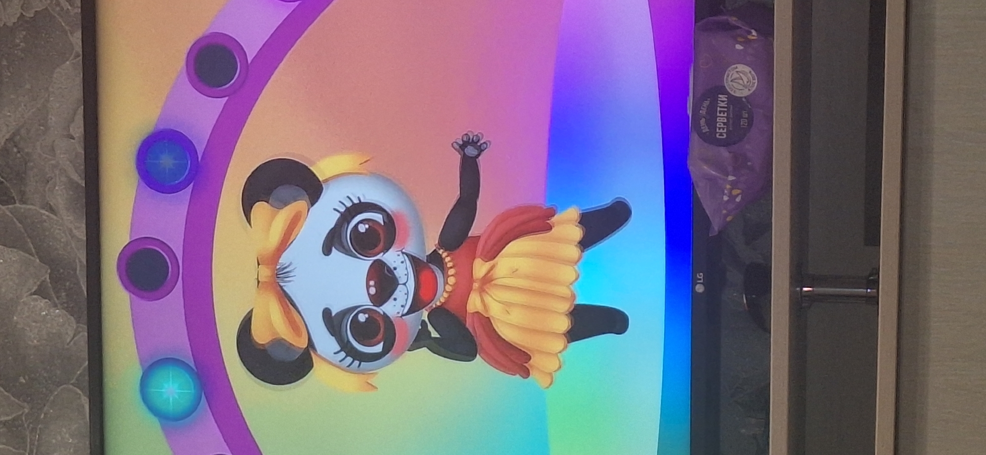 A cartoon panda with large eyes, rosy cheeks, and a yellow bow in its hair, is depicted on a screen. The panda is wearing a red and yellow dress and a pearl necklace. It stands against a colorful background that resembles a rainbow. In the foreground, on a shelf below the screen, a purple package of wet wipes is visible. The LG logo is present on the bottom edge of the screen. The scene appears to be indoors, possibly in a home, given the presence of the screen and shelf. There is no discernible information linking this image to Bila Tserkva, Ukraine.