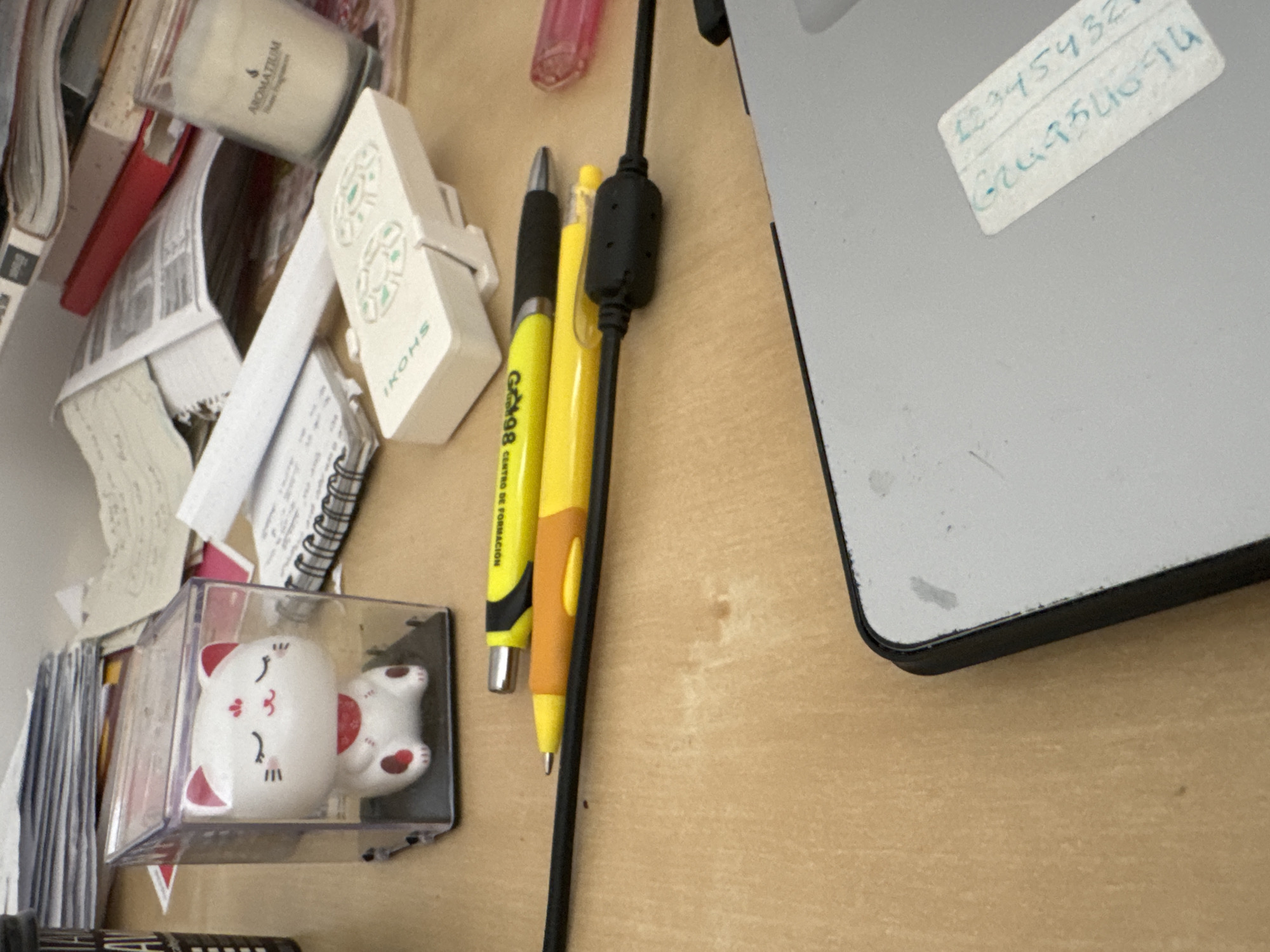 The image depicts a cluttered desk surface, likely in an office or study environment. A white cat figurine in a clear plastic case is prominently featured. Two pens, one yellow and one orange, lie horizontally on the desk, alongside a black cord. In the background, stacks of papers, a red book, and a lit candle in a glass jar are visible. A white remote control with green buttons is also present. The lower right portion of the image shows a portion of a laptop or similar electronic device with a white label attached, bearing handwritten alphanumeric characters. The surface of the desk appears to be wood-grain laminate. The image lacks any specific landmarks or context that would allow for precise geolocation within Murcia, Spain.