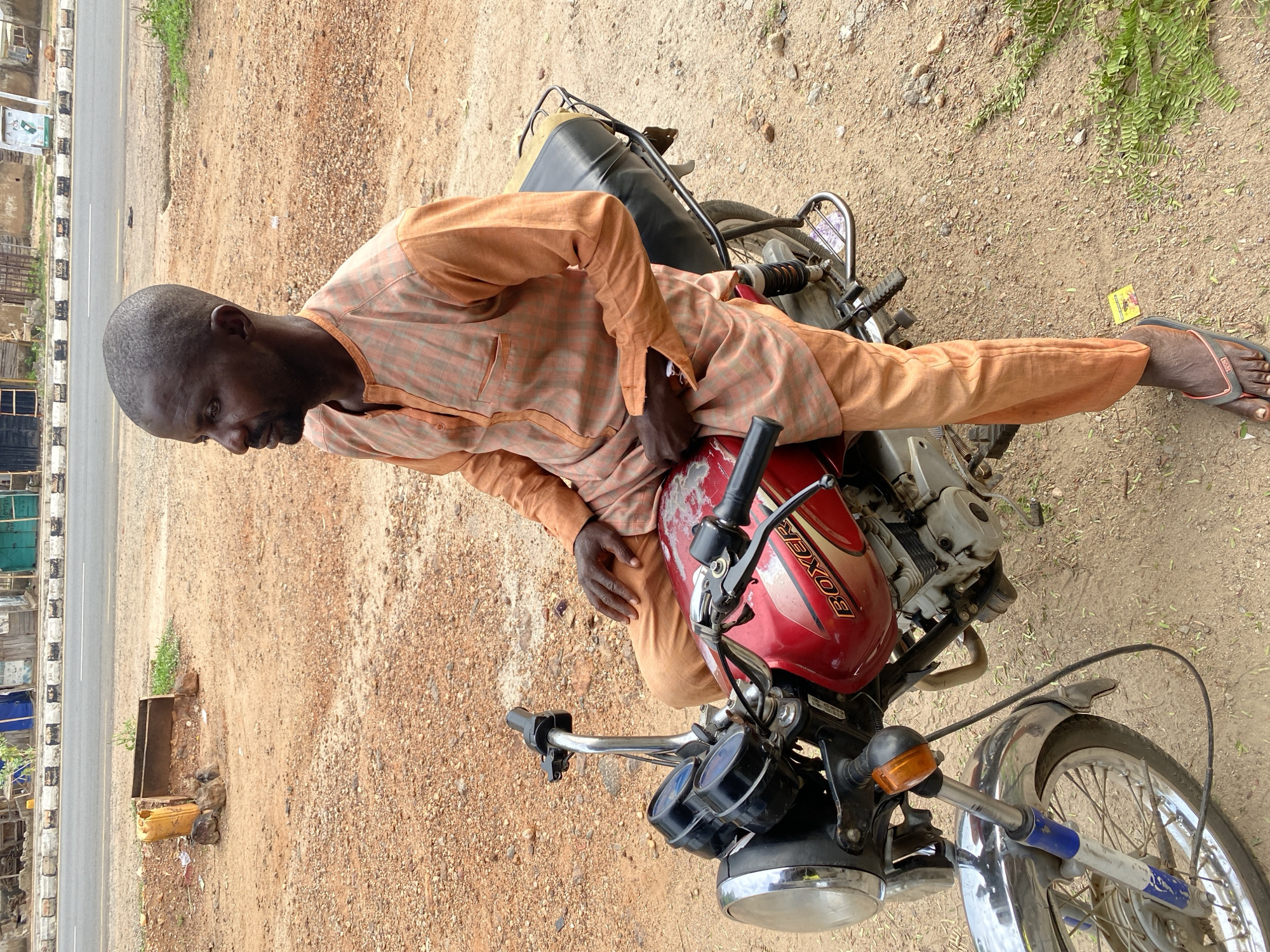 An eye-level, portrait-oriented photograph captures a man sitting on a red motorcycle on a dirt shoulder next to a paved road in Adankolo, Nigeria, during the daytime.
The central subject is a dark-skinned man, appearing to be middle-aged, with a shaved head and a short beard. He is dressed in a light orange plaid-patterned long-sleeved tunic and matching plain orange trousers. He wears black flip-flops on his feet. The man is looking downwards, towards the ground or the motorcycle's front.
He is seated on a red motorcycle with a black seat. The word "BOXER" is visibly written in white letters on the motorcycle's fuel tank. The motorcycle features chrome details on its front fender, handlebars, and headlight. The man's hands rest near the handlebars or on the tank.
The setting consists of a sandy brown dirt shoulder, interspersed with small pebbles, where the motorcycle is parked. To the left of the dirt, a paved road is visible with white lines marking its edge, and a black and white curb. Beyond the road, some rudimentary structures, possibly shops or homes, can be seen in the distance. Sparse green vegetation grows along the dirt and in the background. The bright, natural lighting indicates it is daytime. On the ground near the man's right foot, a small yellow packet with red and black text is visible, though the text is illegible. The scene suggests a casual roadside moment, possibly a wait or a rest.
