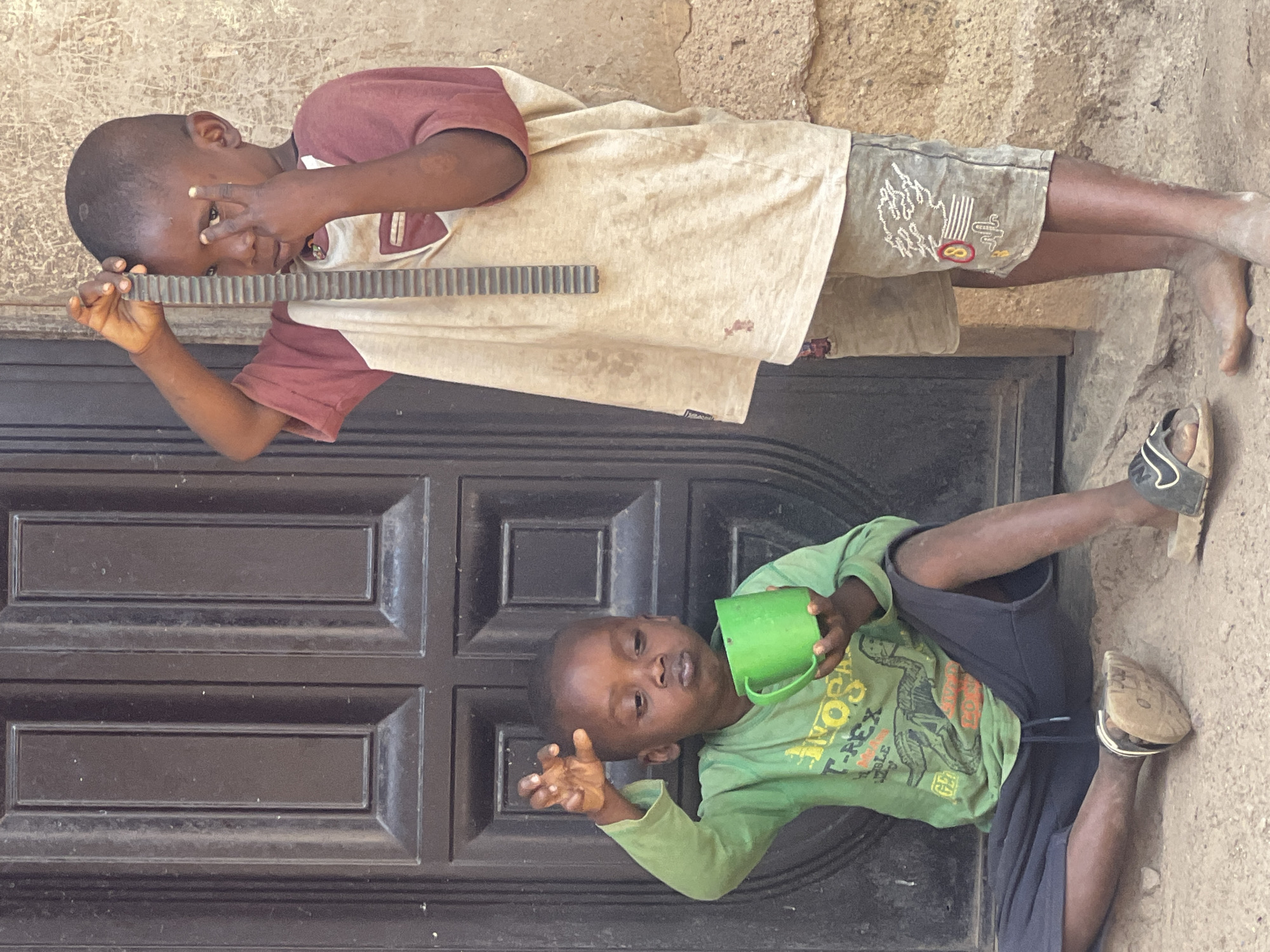 Two young children are positioned in front of a dark, multi-paneled door. The first child, wearing a maroon and cream-colored shirt and patterned shorts, holds a black, comb-like object up to their eye, making a hand gesture. The second child, lying on the ground, wears a green shirt and dark shorts, holding a green cup in one hand and making a gesture with the other. The location appears to be outdoors, possibly near a building made of rough, tan-colored material. The image was taken in Gura Top, Nigeria.
