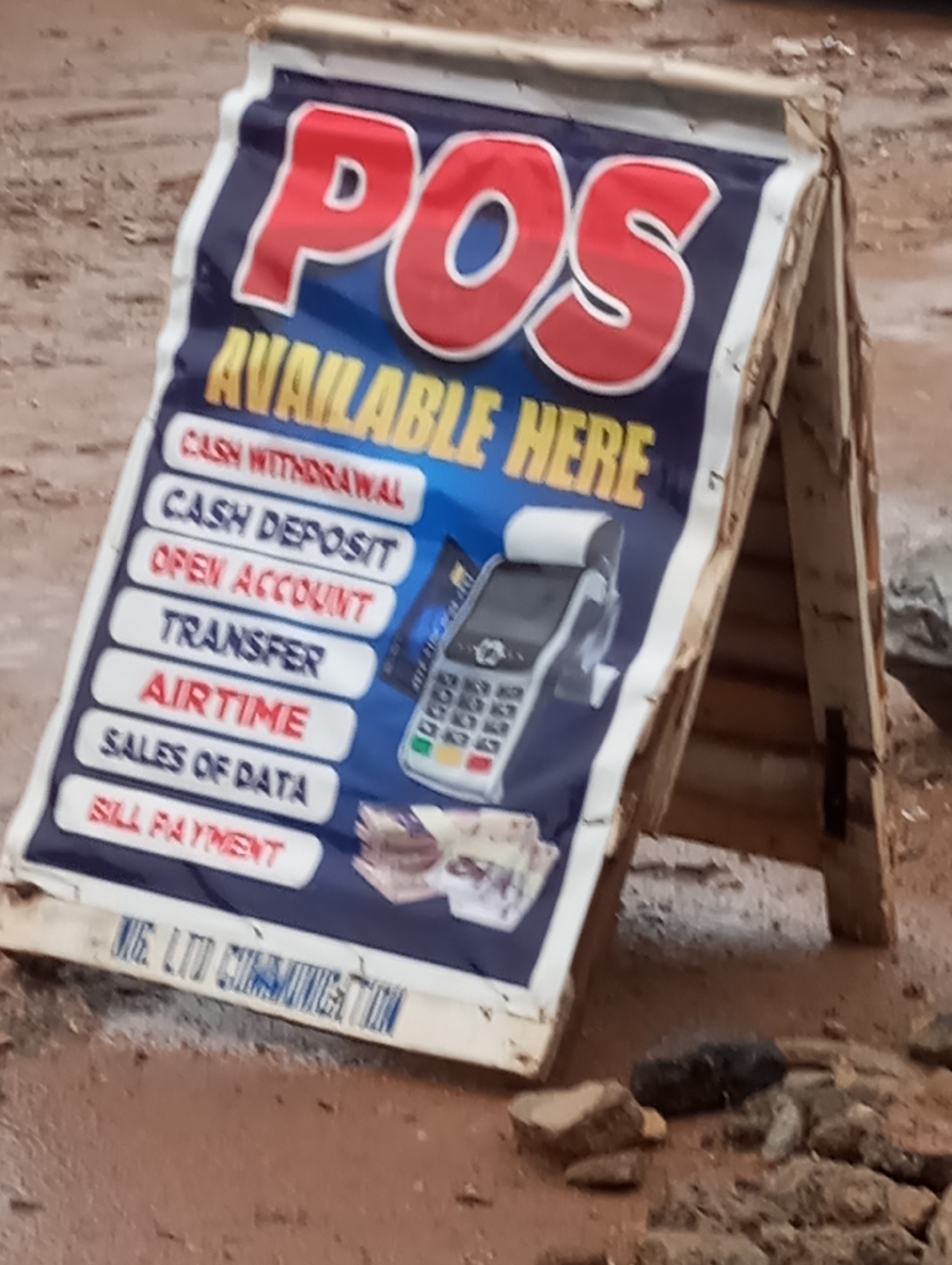 The image shows a weathered sign advertising POS (Point of Sale) services in Sabon Tasha, Nigeria. The sign, propped up on a wooden A-frame, lists available services such as cash withdrawal, cash deposit, opening an account, transfer, airtime, sales of data, and bill payment. A POS machine and a credit card are pictured on the sign alongside some money. The bottom of the sign says "BIG. LTD COMMUNICATION." The surrounding ground is muddy and appears to be a construction site with brick rubble and other debris.