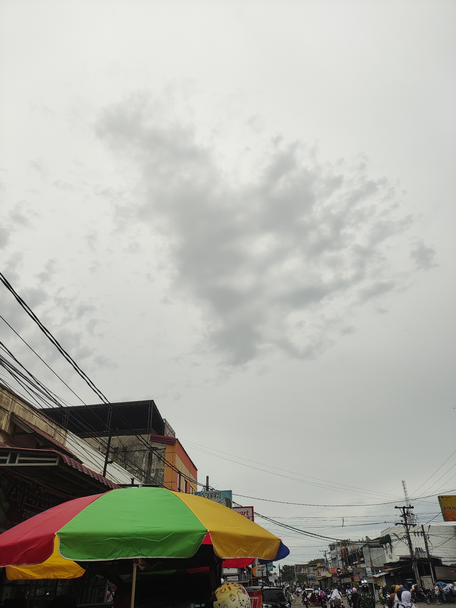 The image depicts a street scene in Lhokseumawe, Indonesia, under an overcast sky. In the foreground, a colorful umbrella with red, green, and yellow panels partially obscures the view. Buildings line the street, and numerous power lines crisscross overhead. A moderate amount of vehicular and pedestrian traffic is present. People on scooters and motorcycles are navigating the street, and pedestrians can be seen walking. Some businesses have signs displayed. The overall scene suggests a bustling urban environment.