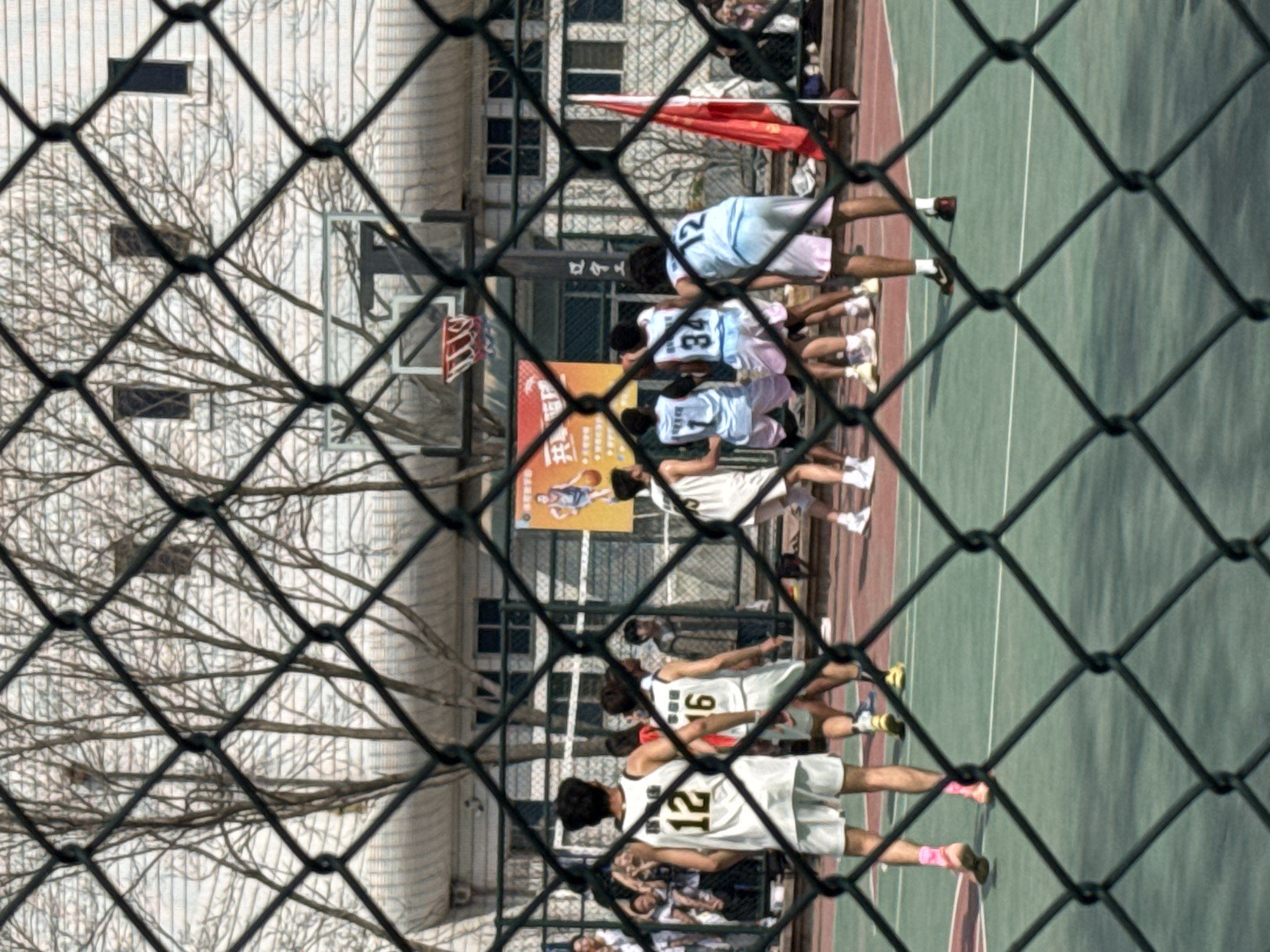 From behind a chain-link fence, the photograph depicts a basketball court in Zhongtun, China. Players wearing white and blue jerseys are visible on the court. There is a building and a leafless tree beyond the court. A red flag is visible above the players.