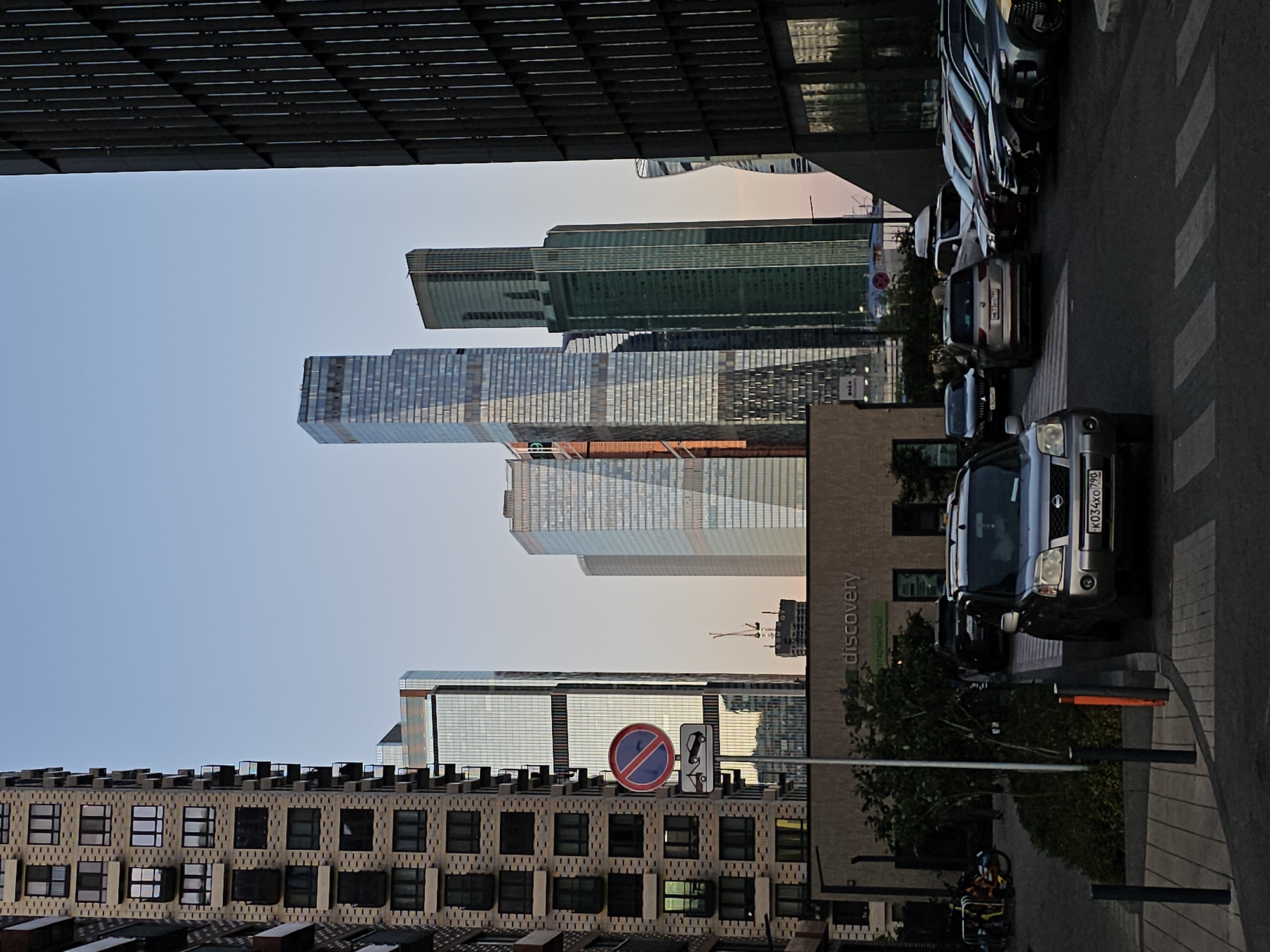 The image depicts a street-level view of a modern urban environment, likely Moscow, Russia, given the architectural style of the skyscrapers and the Cyrillic text visible on signage. The scene is dominated by a collection of towering, contemporary skyscrapers with reflective glass facades, interspersed with a taller, dark-colored building on the right. In the foreground, several cars are parked and driving on a paved road marked with a pedestrian crossing. A silver SUV is prominently featured in the lower-left portion of the frame. A low-rise building with signage reading "discovery" is visible between the foreground and the skyscrapers. There are traffic signs, streetlights, and trees. The sky is clear and appears to be during either sunrise or sunset, casting a warm glow. No people are clearly discernible in the scene, though faint figures might be present within the vehicles or on the sidewalk. The overall impression is one of a bustling, modern city district.