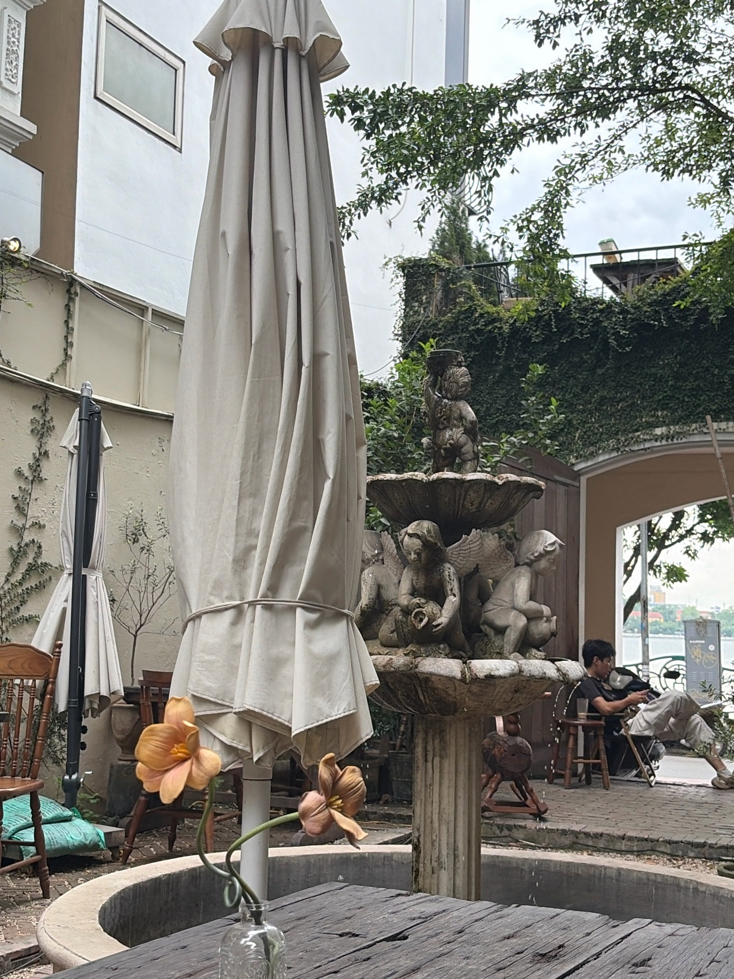 The image captures an outdoor seating area at a cafe in Hanoi, Vietnam. A large, closed umbrella dominates the foreground, obscuring much of the view. To its left, another umbrella is visible, partially furled. In the middle ground, a stone fountain adorned with cherubic figures is the focal point. Beyond the fountain, a man is seated on a chair, looking down at a device in his lap. He is wearing a black t-shirt and light-colored pants. The background reveals lush greenery, including ivy-covered walls and trees. A body of water, likely a lake or river, is visible in the distance, with buildings and the sky beyond. The overall atmosphere suggests a relaxed setting, possibly a courtyard cafe with a view of the water.