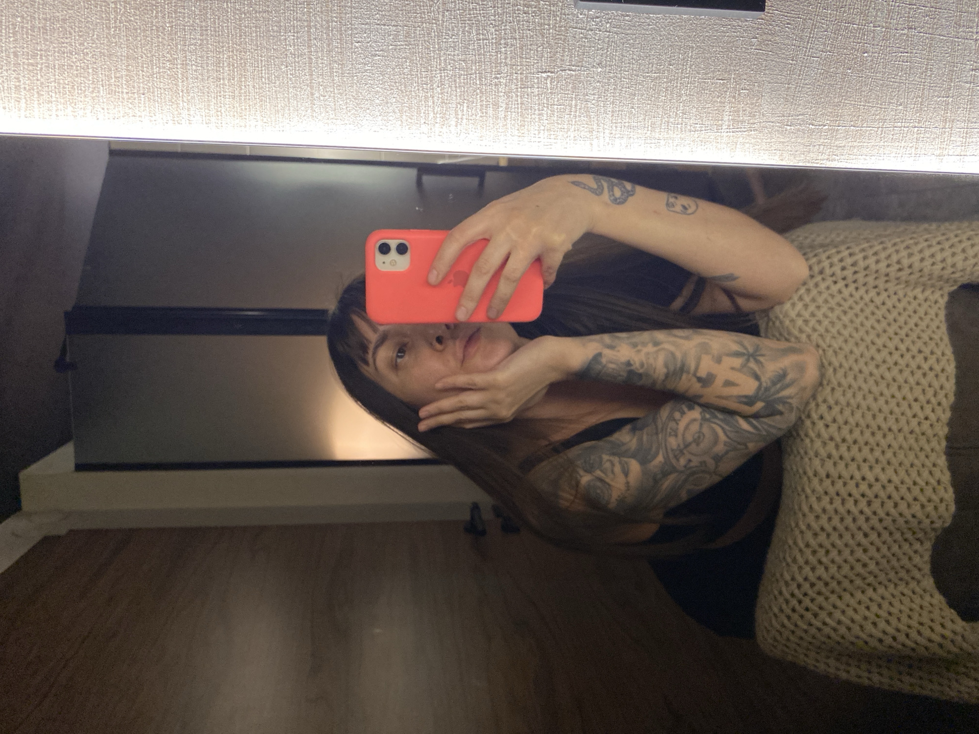 The image captures a mirror selfie taken by a person in an indoor setting, identified as Votkinsk, Russia. The person, a young adult with long, dark hair and bangs, is reflected in what appears to be a large mirror. They have extensive tattoos covering both arms, featuring intricate designs. The person is wearing a dark top and has a light-colored, knitted garment, possibly a blanket or sweater, draped over their left shoulder and arm.
The individual is holding a red-orange smartphone with a dual-camera system visible on the back, suggesting it's an iPhone in a brightly colored case. Their right hand holds the phone, while their left hand is gently touching their cheek. Their expression is calm and neutral.
Above the mirror, a long, linear light fixture illuminates a textured, light-colored wall or panel. Below the mirror, a dark, reflective surface, possibly part of a dresser or shelf, is visible, resting on a lighter-colored base. The floor is made of dark brown wood or laminate. The overall lighting suggests an indoor evening setting or an area with limited natural light, relying on artificial illumination. There is no visible text.