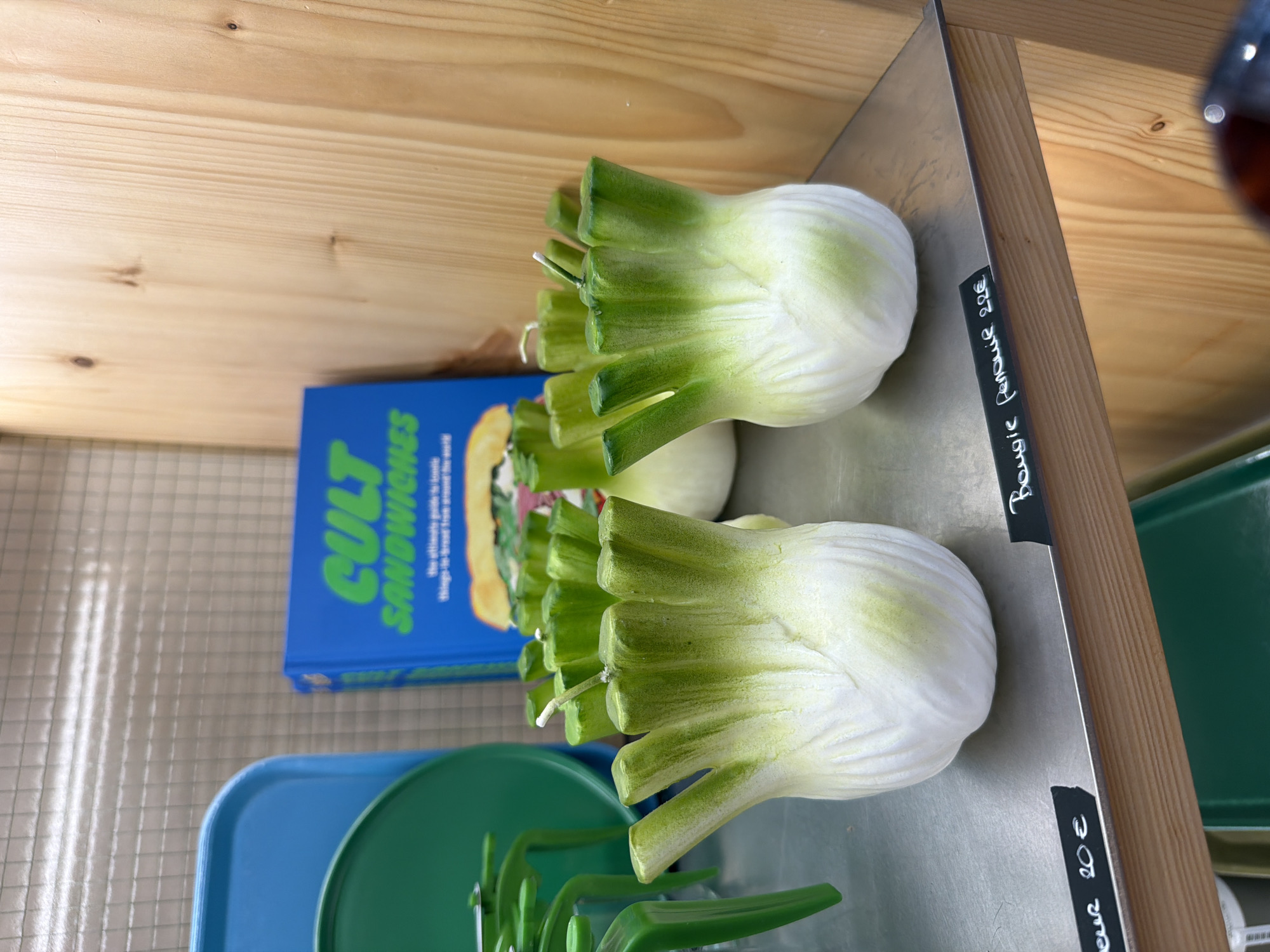 The image shows a close-up of a shelf with two fennel-shaped candles, each with a white bulb and green stalks. The candles have wicks protruding from the top of the stalks. In the background, a blue book titled "CULT SANDWICHES" is visible. To the left of the book, a portion of a blue tray and a green circular object are seen. The shelf appears to be made of metal and is positioned against a wooden wall. There are price tags on the front edge of the shelf; one reads "cur 20€" and another reads "Bougie Fenouil 20€". The lighting suggests an indoor retail environment. The scene does not provide any explicit information about its location in Paris, France, beyond being an indoor retail display of novelty candles and a book.