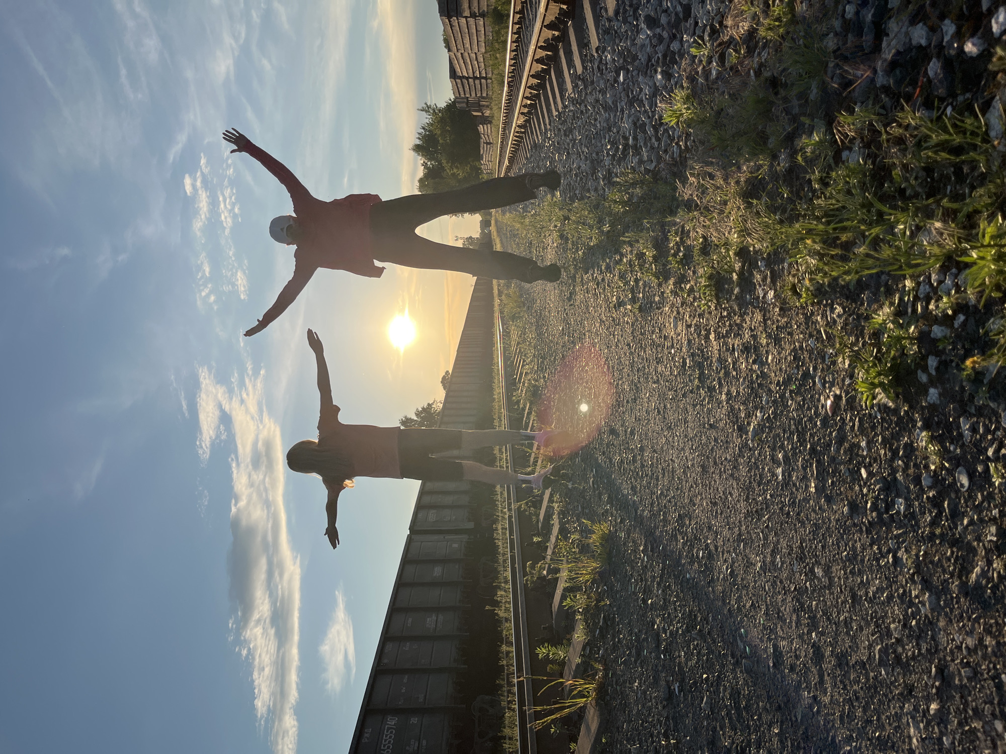 The image shows two people standing on railroad tracks at sunset. One person, a child, is on the left with their arms outstretched to the side. The other person, an adult, is on the right with their arms raised above their head. Both are silhouetted against the bright sun. The railroad tracks stretch into the distance, with freight cars visible on the left side of the frame and some wooden structures on the right. The sky is blue with scattered clouds, and the sun is setting behind the trees in the distance. The ground is covered in gravel and scattered vegetation. The scene appears to be in a rural or industrial area, possibly on the outskirts of Chaikovsky, Russia, given the presence of what appear to be Russian freight cars.