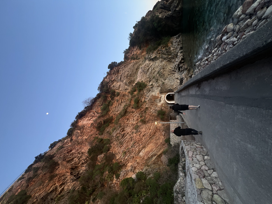 A coastal scene in Rafailovići, Montenegro, depicts a paved pedestrian pathway running alongside a steep, layered reddish-brown cliff face. The cliff is partially covered with sparse green vegetation and is illuminated by warm, low-angle sunlight, indicating either dawn or dusk. To the right of the pathway, the clear, turquoise-colored sea gently laps against a rocky shoreline, separated by a concrete retaining wall.
Ahead on the pathway, a pedestrian tunnel entrance is visible, carved into the cliff face, and appears illuminated from within. Two operational streetlights with a yellow glow are positioned along the pathway. High in the clear, light blue sky above the cliff, a distinct white crescent moon is visible.
Two individuals, both dressed in dark clothing, are walking away from the camera along the pathway towards the tunnel. The person on the left wears long pants and dark shoes. The person on the right wears shorts and appears to have light-colored footwear or is barefoot. Their backs are to the viewer; no facial features are discernible.