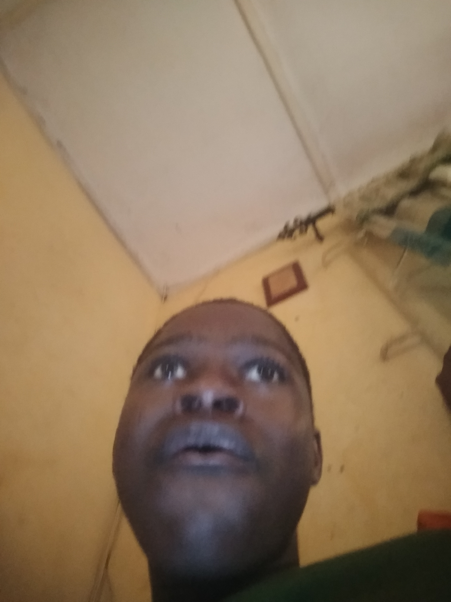 A low-angle, close-up photograph captures a person's face, filling much of the frame. The individual has dark skin, short hair, and is gazing upwards. The background consists of a room with yellowish walls and a white ceiling. An object that appears to be a gun is mounted on the wall, along with a framed picture. The location is Unknown, Unknown.