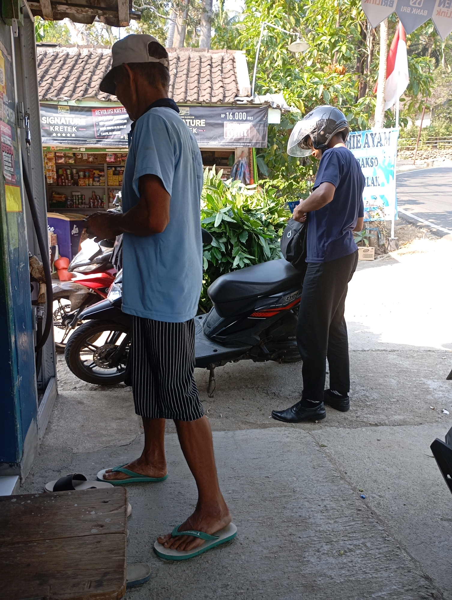 In Ketepung, Indonesia, two men are standing near a small shop and two motorcycles. The man on the left, wearing a light blue t-shirt, striped shorts, and flip-flops, is standing at a gas pump, holding a hose. He has a cap on his head. The man on the right, dressed in a dark blue t-shirt, black pants, and dress shoes, is wearing a motorcycle helmet with the visor up and is holding a bag. A black motorcycle is parked behind him, and a red motorcycle is partially visible to his left. The shop appears to sell various goods, with shelves of items visible inside. The setting is outdoors, with trees and foliage in the background, and the ground is paved.