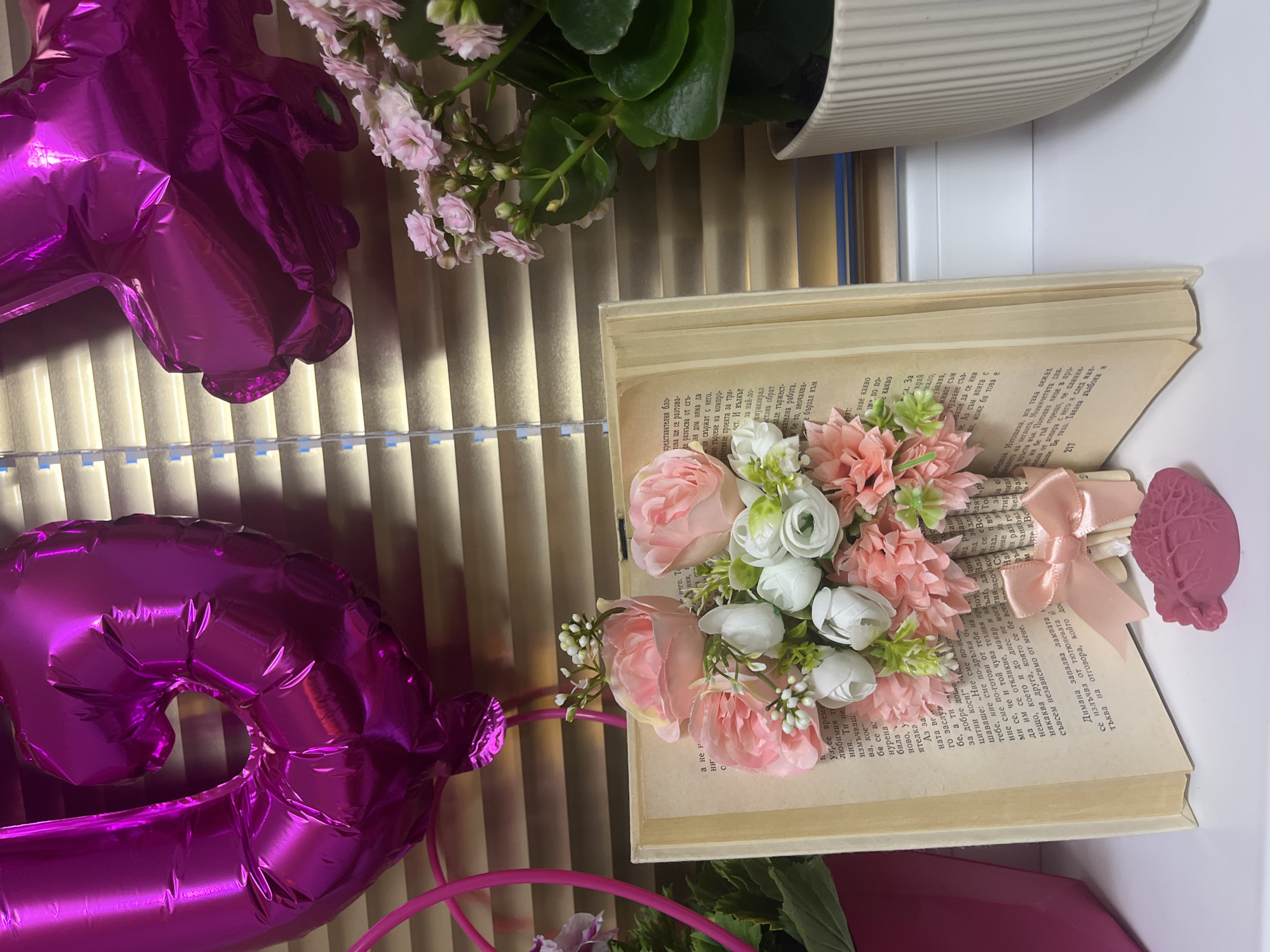 This is a still life arrangement. There is a vintage book lying open, with pink and white flowers arranged on its pages. Beside the book are pink balloons, and green plants, and in the background are blinds.
There is no indication this image was taken in Asenovgrad, Bulgaria.
