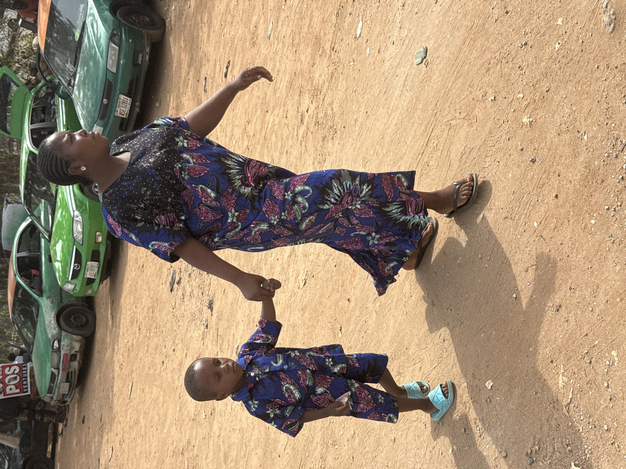 A woman and a young child are standing in a sandy area next to a row of green cars. The woman is wearing a blue dress with a floral pattern and black top. The child is wearing a matching blue floral shirt and shorts, with blue sandals. They are holding hands. The area is sandy and dusty. Cars are parked nearby. A red sign reading "POS VENTURES" is visible on one of the cars. The city of Abuja, Nigeria, cannot be identified from the provided information.