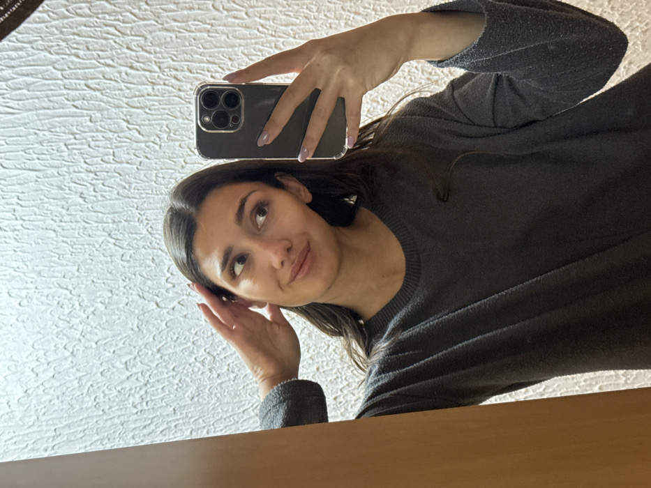 The image displays an indoor mirror selfie of an adult female located in Zankovići, Montenegro. The female subject has long, dark brown hair and is wearing a dark gray, long-sleeved textured top. Her right hand is positioned against the right side of her head, with fingers splayed. Her left hand holds a dark-colored smartphone, equipped with a clear protective case and a visible multi-lens camera array, positioned to capture the reflection. The subject's gaze is directed towards her right, away from the camera, and her lips form a slight smirk. Both hands display fingernails painted with a light-colored polish. The background behind the subject's reflection features a light-colored wall with an irregular, raised textured pattern, and a vertical, light brown wooden architectural element is partially visible on the far left. The scene is illuminated by general indoor lighting.