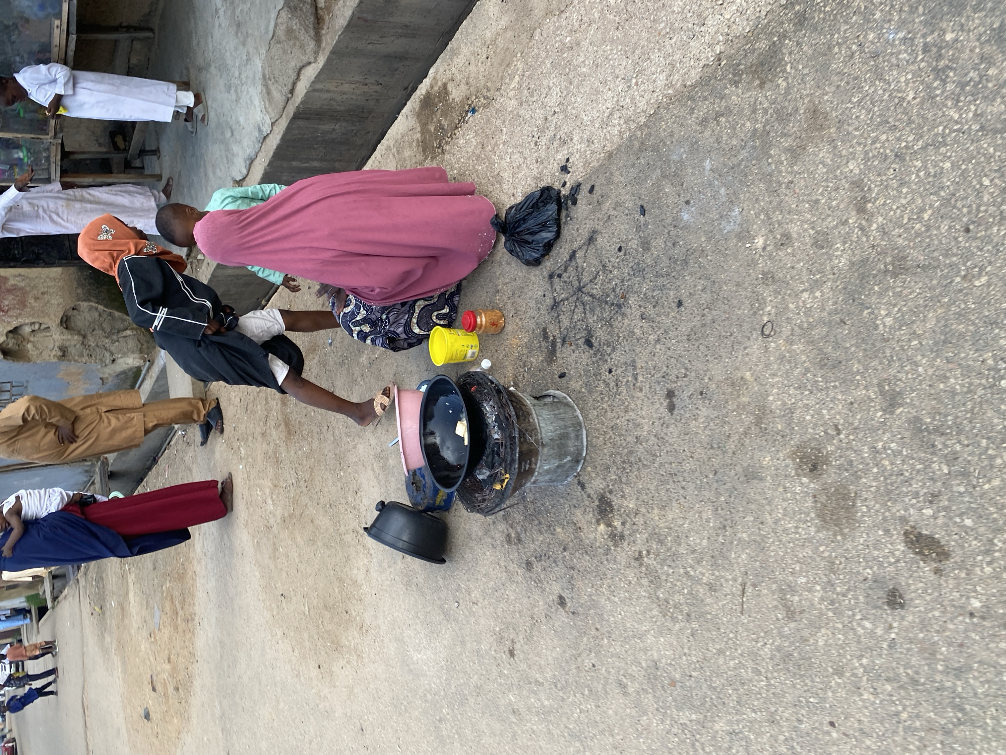 In Jos, Nigeria, on a paved street, a group of people are gathered. In the foreground, a metal charcoal stove with burning embers is being used for cooking. A black pot sits on the stove, containing food. To the left of the stove is a black plastic basin, and to the right, a pink bowl. Nearby, a yellow container and a small jar with red spices are placed on the ground. A black plastic bag lies on the ground in front of a person wearing a pink hijab, who is seated with another person dressed in a patterned fabric. Behind them, another child in a green garment is visible. Further in the background, a person in a black and orange hijab bends over, and a child in a white garment stands near a wooden table. Other individuals in traditional attire are also present on the street, with some buildings and a gutter in the background.