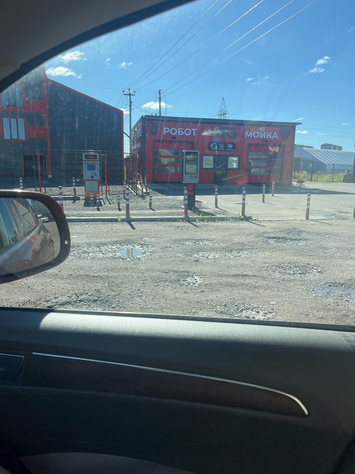 The image depicts a car wash facility under a bright, sunny sky with scattered clouds. The car wash building is red with "РОБОТ" and "МОЙКА" (Robot and Car Wash in Russian) written on its facade. A payment kiosk is visible in front of the building, along with several bollards. To the left, another building with dark gray siding and red accents is partially visible. The ground in front of the car wash is unpaved and appears to be gravel with some puddles. In the foreground, a portion of a car's interior, including the driver's side door panel and rearview mirror, is visible, suggesting the photo was taken from inside a vehicle. The overall scene suggests an outdoor commercial area. It is difficult to pinpoint the exact location within Saint Petersburg without further context or identifying landmarks.