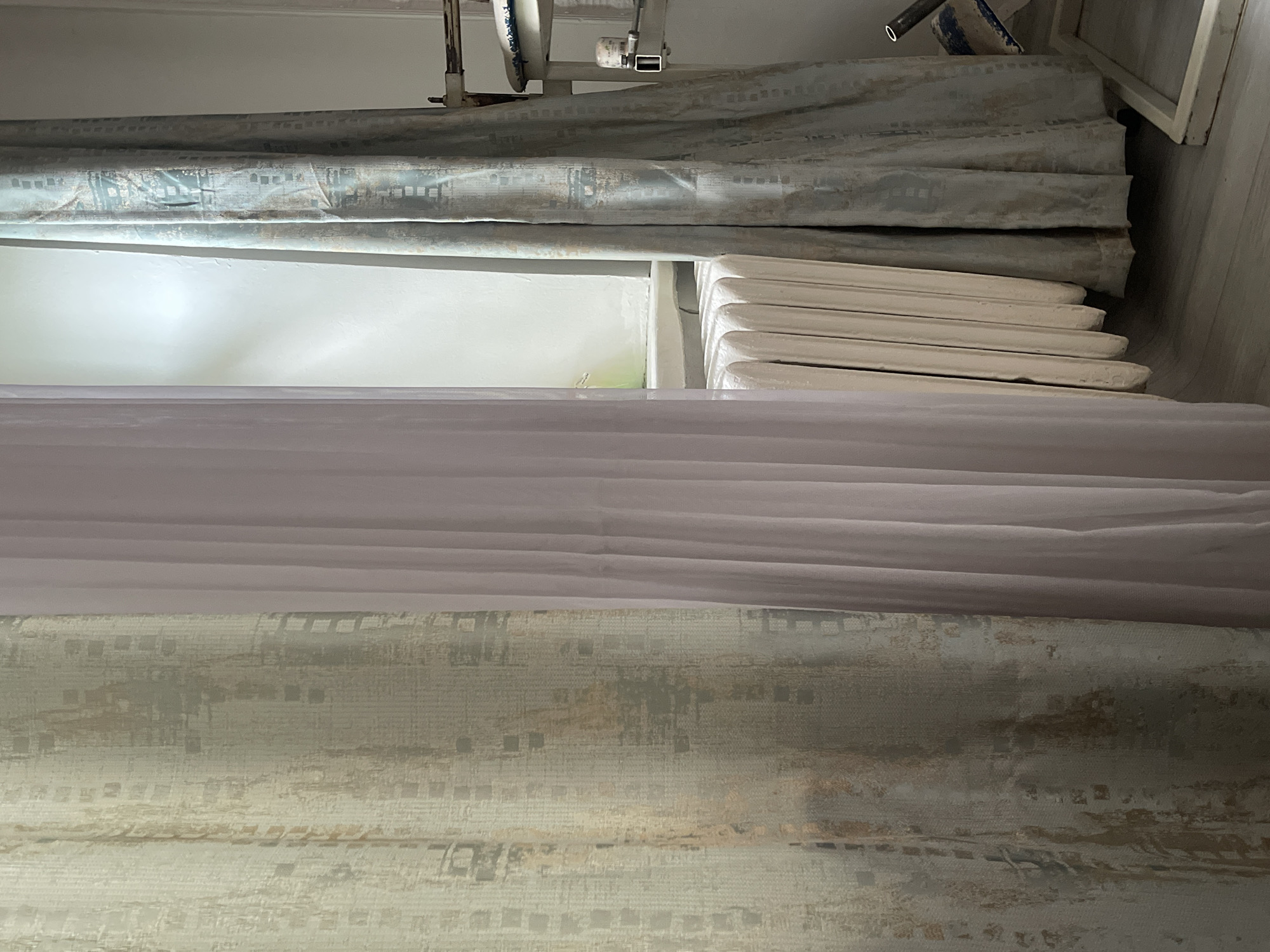 The image depicts a close-up view of a window area in an indoor setting, likely a room. Three curtains are visible: a light-colored, textured curtain on the left, a sheer, pale pink curtain in the center, and a patterned, muted blue-green curtain on the right. Behind the sheer curtain, a white radiator with multiple fins is situated beneath the window. The window itself is partially obscured by the curtains, but light is filtering through, illuminating the wall behind the left curtain and creating a bright spot on the wall above the radiator. The flooring appears to be light-colored wood or laminate. To the far right, part of a white metal frame structure, possibly furniture or an appliance, is visible, with a white object and blue fabric attached to it. The scene is static and does not show any people or ongoing actions. The location is not specified and cannot be determined from the image alone.