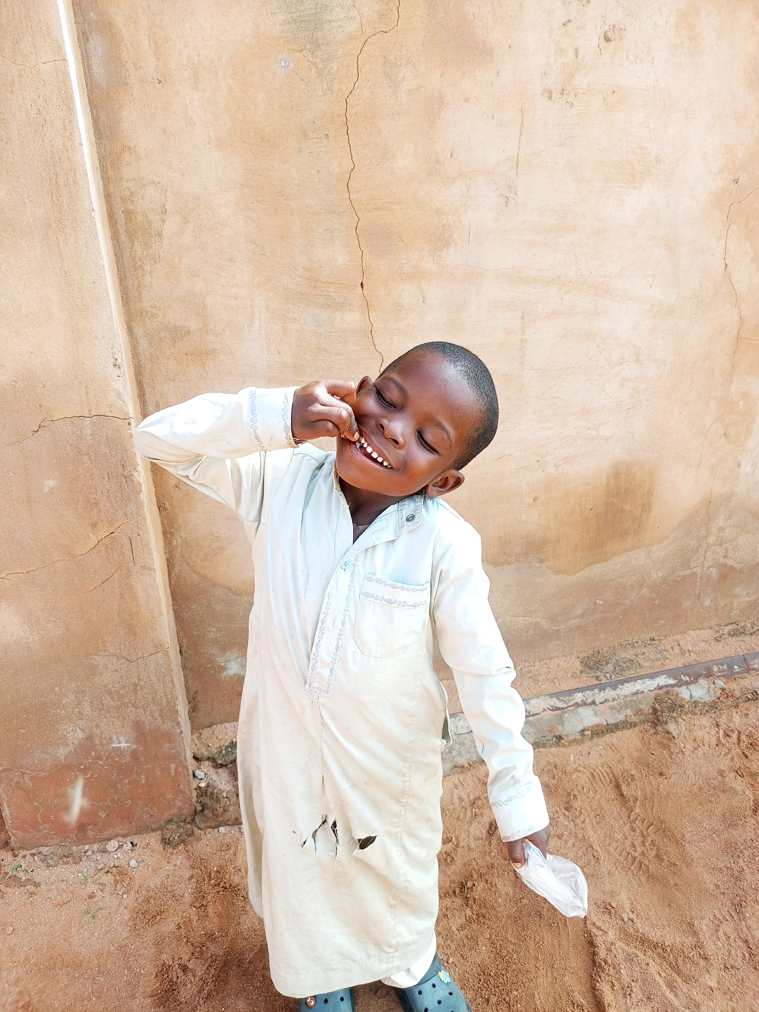 A young Black boy, appearing to be around 5-7 years old, stands in front of a textured, beige-colored wall, possibly made of mud or stucco. The wall has visible cracks and some discoloration, suggesting it is aged. The ground beneath the boy is sandy and dusty, with some small stones and dried vegetation scattered about. The boy is wearing a loose-fitting, light-colored robe, possibly a traditional garment, which has several large holes torn in the lower front portion. On his feet, he wears dark-colored sandals.
The boy is captured mid-action, with his eyes closed and a wide smile on his face, his head tilted slightly to his right. His left hand is raised, and his index finger is touching his cheek near his mouth, giving the impression that he is making a playful or teasing gesture. In his right hand, he holds a crumpled piece of white material, possibly plastic or paper.
The scene appears to be outdoors, likely in a somewhat informal or rural setting within the city of Tempure, Nigeria. The lighting suggests it is daytime, with the sun casting subtle shadows.