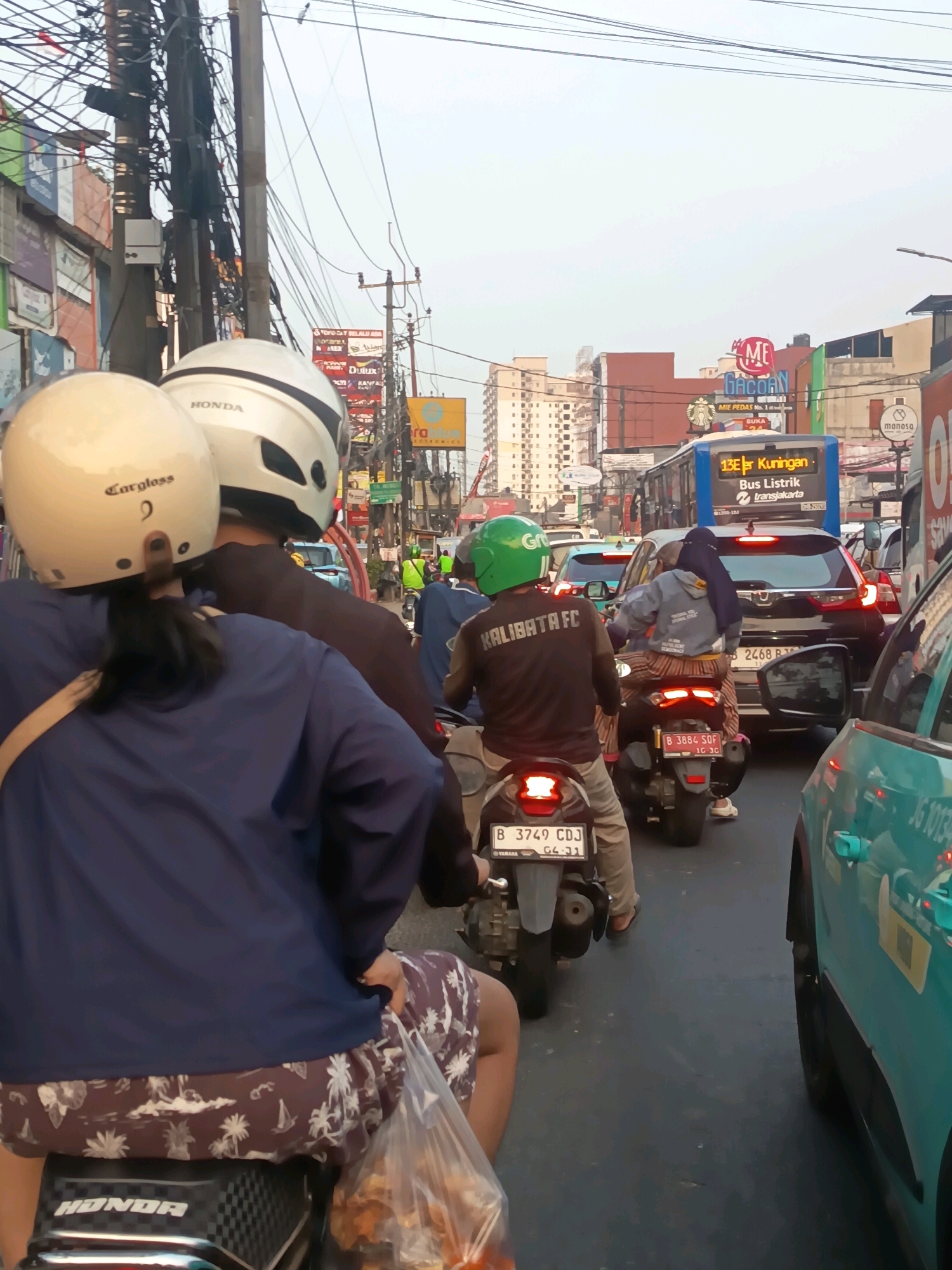 A street scene in Tangerang, Indonesia, depicts moderate traffic congestion, primarily consisting of motorcycles and cars. In the foreground, two individuals wearing helmets are riding a motorcycle. The person in the rear is wearing a blue jacket and patterned shorts, while the person in front has a white helmet with black trim and the brand "HONDA" visible. A plastic bag filled with items is visible on the rear of the motorcycle. The environment shows a busy street lined with buildings, advertisements, and numerous overhead electrical wires. A blue bus with "13E Kuningan" and "Bus Listrik" displayed on its front is visible further down the street. The overall atmosphere suggests a typical urban day in Indonesia with active street life and transportation.