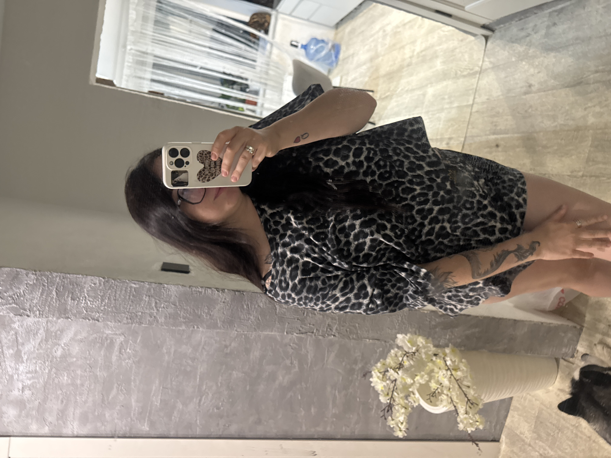 A woman is captured in a mirror selfie, standing indoors. She holds a white smartphone with a black camera array and a leopard-print heart sticker on the back, partially obscuring her face. She has dark hair, wears glasses, and a ring on her left ring finger. She is dressed in a black and brown leopard-print garment. Tattoos are visible on both forearms: a small red design (possibly two drops or hearts) on her left, and a larger, dark tattoo resembling a winged creature (perhaps a bird or phoenix) on her right.
The reflected background reveals an interior space, likely a home. To the left, a textured grey wall is visible. The floor is covered with light-colored, possibly beige or cream, tiles. In the lower right of the reflection, a tall, white ribbed vase filled with white flowers on branches is positioned. Below this, a portion of a dark-furred animal, resembling a dog, is visible. Further back in the reflection, a window with horizontal blinds allows daylight into the room. A blue plastic water bottle and a light-colored chair are also faintly visible in the distance of the reflection. The overall impression is an everyday moment, likely taken during the daytime. No clearly legible text is present, although small decorative text is part of the phone case sticker.
