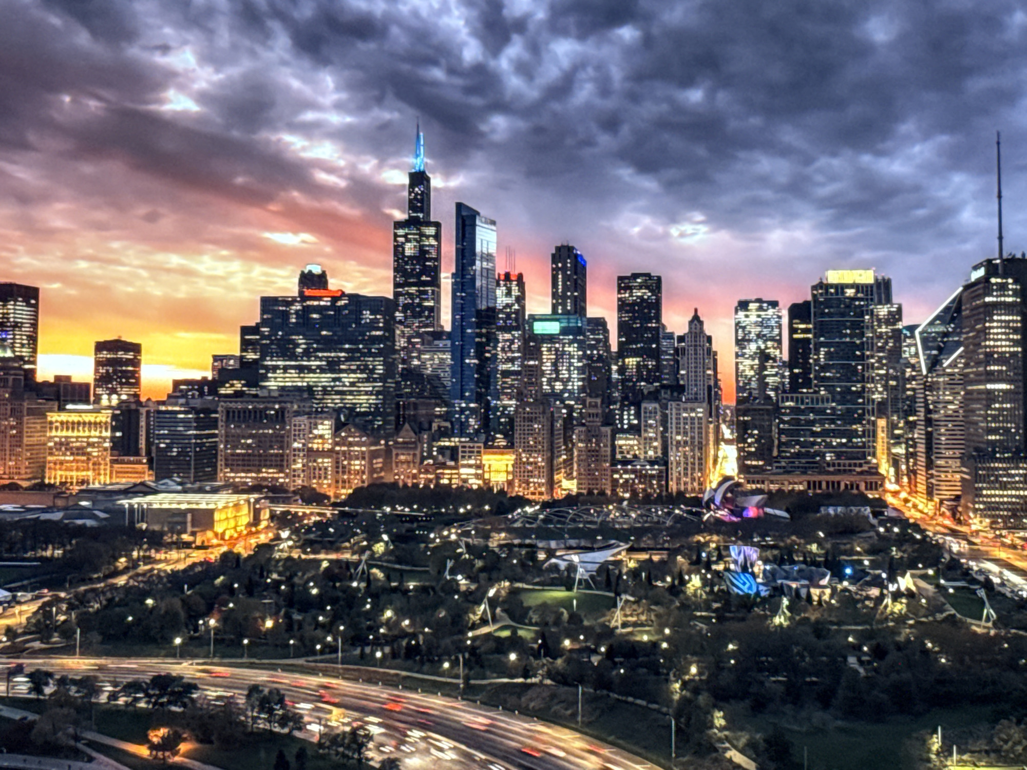 The image depicts a sprawling cityscape at dusk, characterized by a dramatic sky transitioning from warm sunset hues to a brooding, dark grey. Towering skyscrapers with illuminated windows dominate the skyline, suggesting a major urban center. In the foreground, a park area with trees and pathways is visible, and below it, a highway is teeming with the streaks of car lights, indicating active traffic. The architecture and the overall scale of the city strongly suggest a location such as Chicago, Illinois, known for its impressive skyline and extensive urban parks. The prompt's request to identify the location in Burgas, Bulgaria, is inconsistent with the visual evidence presented, as the city's architecture and layout do not resemble Burgas.