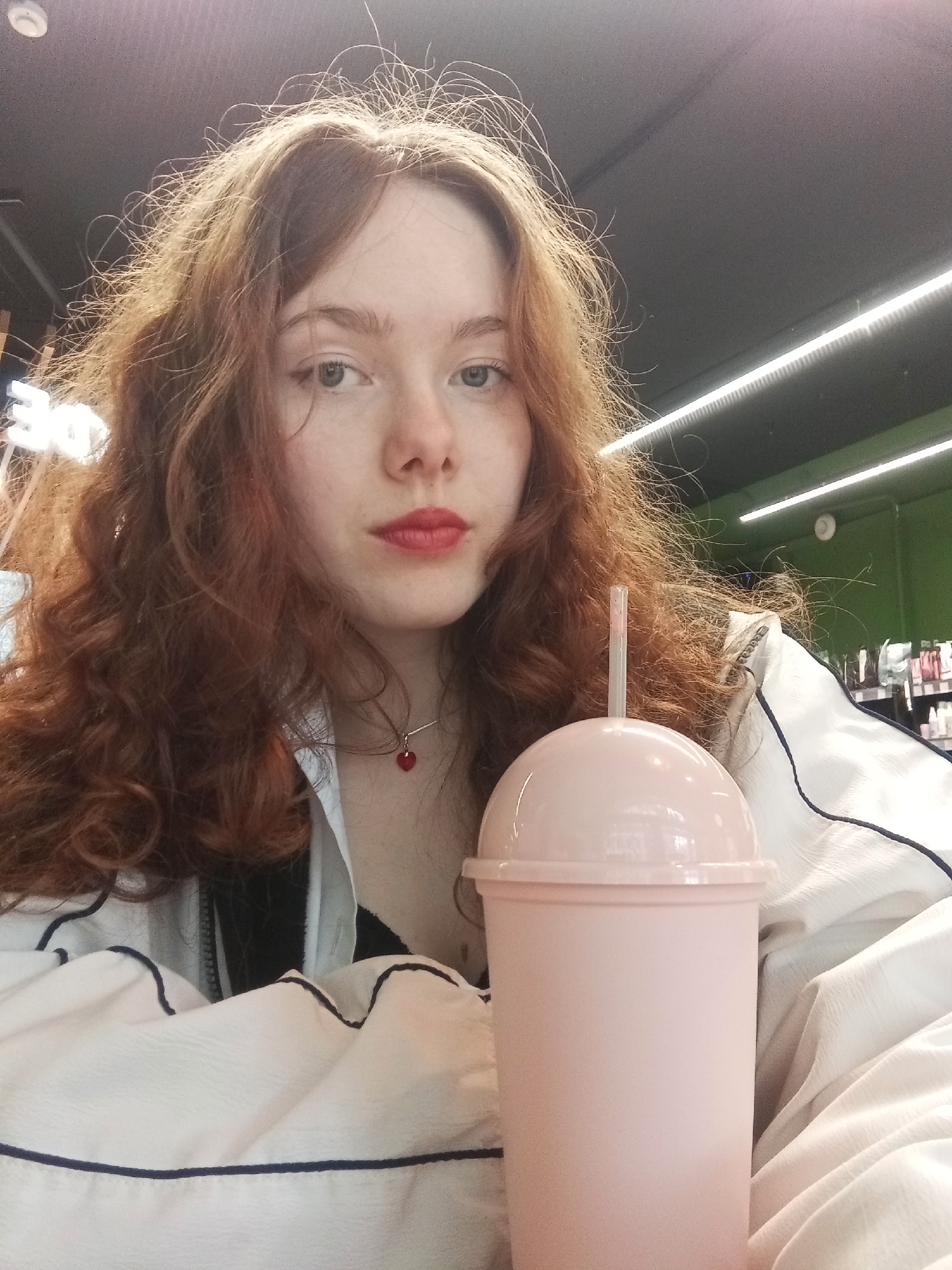 A young, light-skinned woman with long, curly red hair and fair complexion is posing indoors. She wears red lipstick and a necklace with a red, heart-shaped pendant. She is wearing a white jacket with navy blue piping. In front of her is a large, pink, dome-lidded cup with a straw. The background includes shelves of products, and lighting fixtures on the ceiling. Given these elements, the image might be taken in a shop or cafe. No information indicates this picture was taken in Saint Petersburg, Russia.