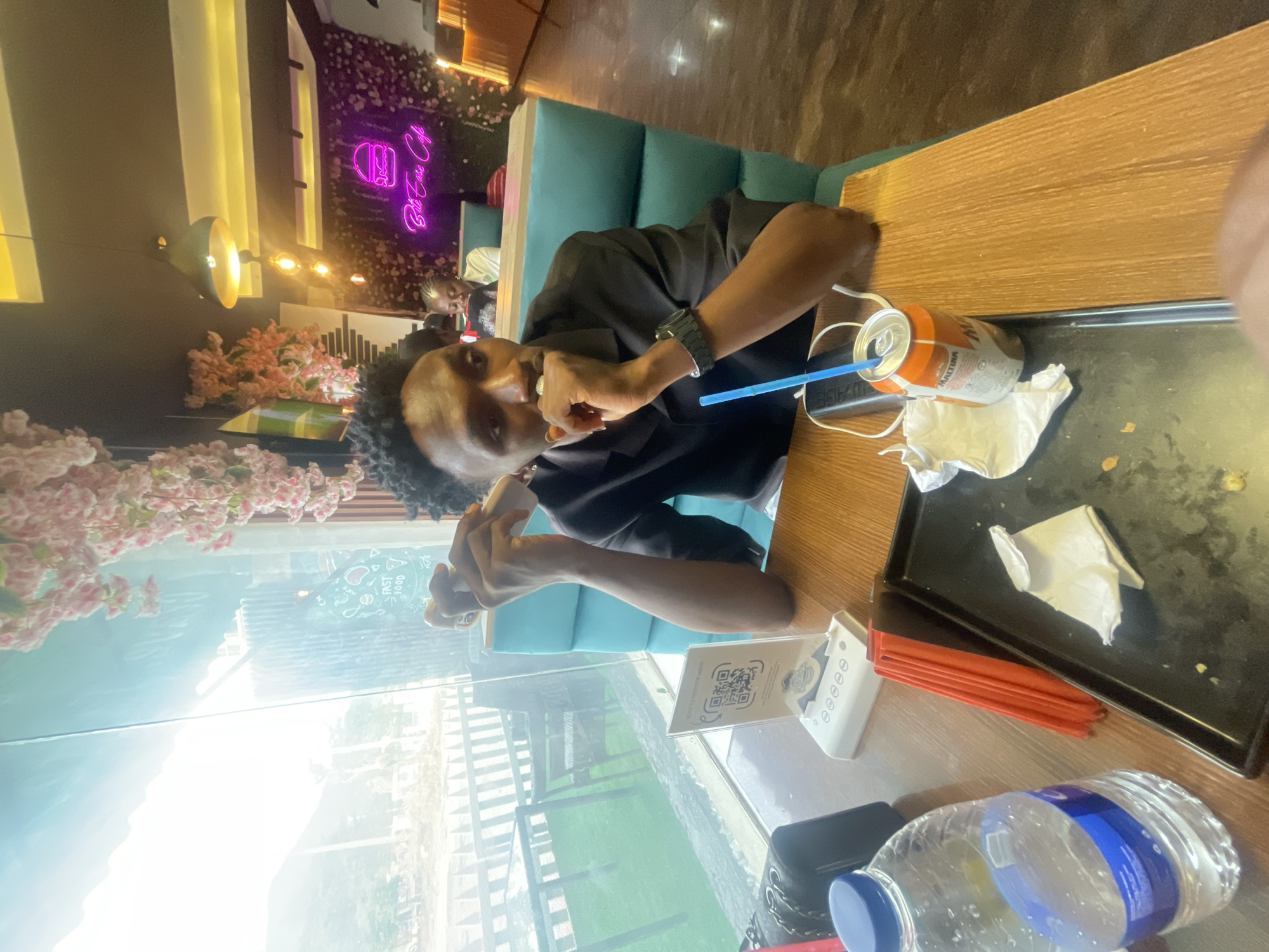 The image captures a young man seated at a wooden table inside a cafe or restaurant in Adankolo, Nigeria. The image is taken during the daytime, with bright natural light streaming in through a large window.
The main subject is a young man wearing a black short-sleeved shirt and a black watch on his left wrist. He has short, dark, curly hair and is looking directly at the camera with a neutral expression. His right hand is raised near his mouth, seemingly holding a small item or piece of food, while his left arm rests on the table.
The table in front of him is cluttered with various items. To his right, there is an orange and white can with a blue straw, identifiable by the text "MALTINA" on its label. A crumpled white napkin is beside the can. Further along the table, a black rectangular tray holds several red straws and another white napkin. A clear plastic water bottle with a blue cap and label rests closer to the foreground. A white stand displaying a QR code with the text "SCAN TO VIEW MENU" is also visible, along with a dark-colored smartphone.
The setting appears to be a modern cafe with teal-colored booth seating. The background features a decorative wall adorned with pink artificial cherry blossom-like flowers. A prominent purple neon sign on this wall displays a burger icon and the text "Butcher Cafe." Modern light fixtures with warm-toned bulbs are also visible in the upper left background. Beyond the glass window behind the man, bright sunlight creates a glare, but glimpses of an outdoor area with greenery and a fence or railing can be seen. Reflections on the window show faint text "FAST FOOD" and various food-related icons. Another person is partially visible sitting further down the booth in the background.