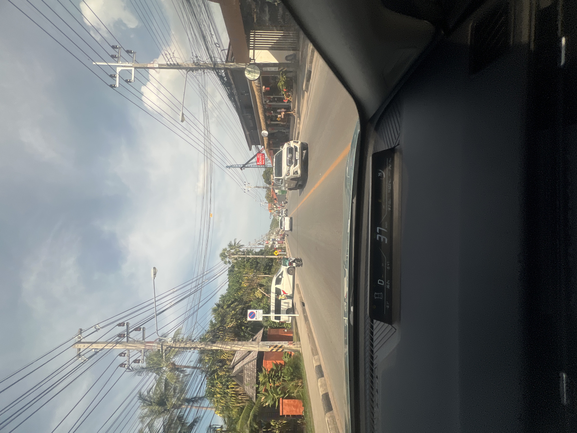A car's perspective shows a street scene in Ko Samui District, Thailand, on a partly cloudy day. Power lines crisscross the sky, supported by utility poles along the road. On the left, a building with a wooden roof and orange walls is visible, alongside palm trees and dense vegetation. A white van with a person standing near the driver's side is parked on the side of the road, next to a "no parking" sign. A motorcycle is also present on the road. The street itself has a yellow dividing line and is lined with shops and businesses on the right side. Several cars are driving on the road, including a white SUV. The car's dashboard displays information, including the current speed of 37 km/h.