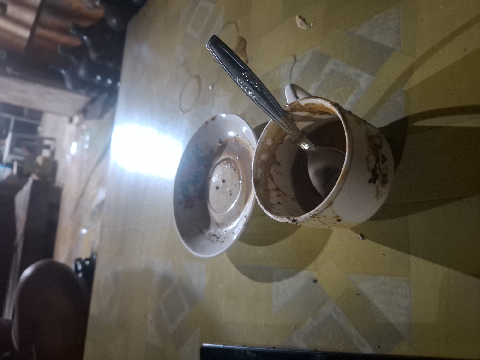 An overhead shot captures a close-up of a coffee cup and saucer on a patterned table. The saucer, positioned to the left, contains a residue of coffee, with a light-colored floral design visible on its rim. To the right, a white ceramic cup, also decorated with floral motifs, is filled with coffee and holds a spoon. The cup's rim shows some spillage. The table surface is yellow with a repeating grid pattern of lighter squares and diamonds. Shadows cast by the cup and saucer stretch across the table. In the blurred background, dimly lit furniture and several large dark bottles are visible. The scene appears to be set indoors, possibly in Bulubrangsi, Indonesia.
