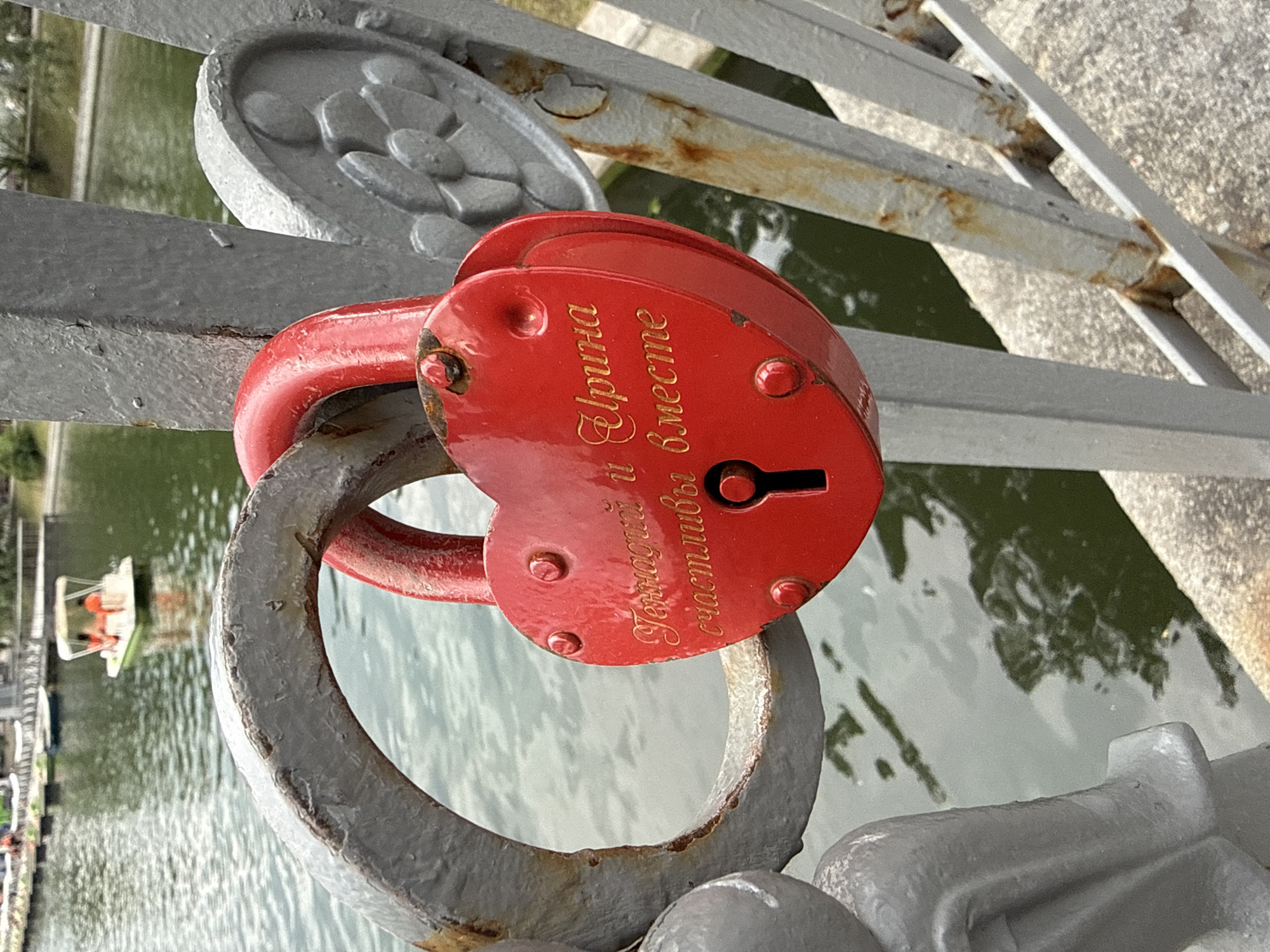 The image captures a close-up of a bright red heart-shaped padlock attached to a weathered gray railing. The padlock is inscribed with golden Cyrillic lettering, which appears to read "Геннадий и Ирина счастливы вместе" (Gennady and Irina are happy together). The railing shows signs of rust and age, contrasting with the vibrant padlock.
In the background, a body of water, likely a canal or river, is visible. A small white boat with two people wearing orange life vests is moving across the water. The far bank of the water features greenery and some indistinct structures, possibly part of an urban park.
This scene is typical of "love locks" found on bridges in many cities worldwide, including Moscow. Specifically, the iconic location for such locks in Moscow is the Luzhkov Bridge, which connects the Moscow State University and the Park of the Great Patriotic War. The railing in the image is consistent with the railings found on this bridge, which are adorned with numerous love locks. The presence of the boat on the water further suggests a recreational area within the city, aligning with the setting of Luzhkov Bridge and its surrounding park.