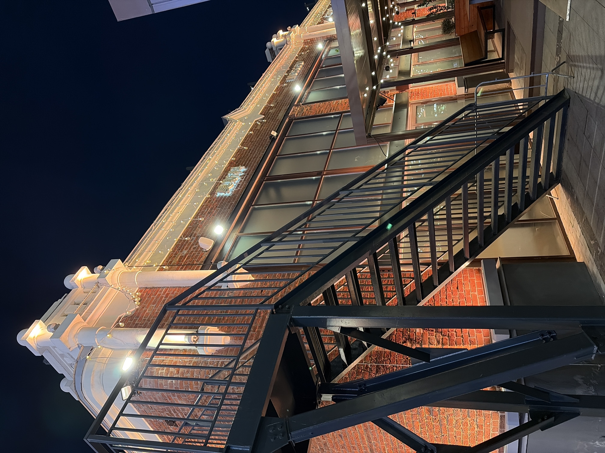 An exterior shot of a brick building at night features an angled view of a black metal staircase in the foreground, leading up towards the building's facade. The building has large windows and is illuminated by strings of warm white lights and spotlights that cast an orange glow on the brickwork. White decorative elements adorn the top of the building. The scene appears to be an outdoor seating area for a restaurant or bar, with some seating visible under a covered area to the right. The sky is dark blue, indicating nighttime. The specific location in Moscow, Russia is not identifiable from the visual information provided.