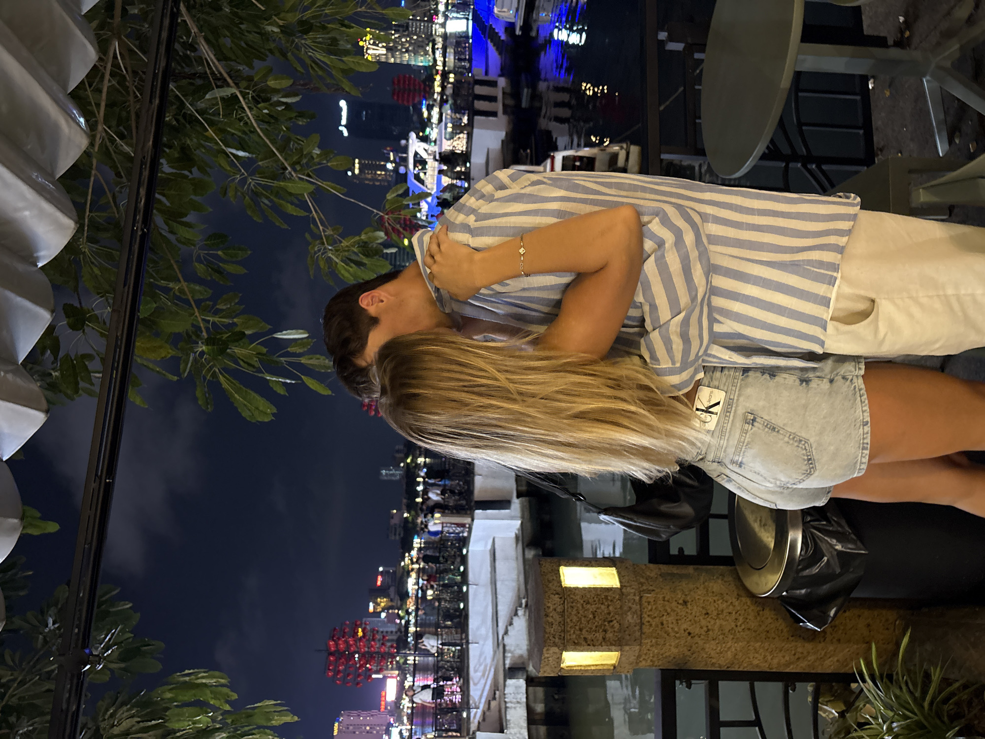 A couple kisses outdoors at night. The man wears a blue and white striped shirt and white pants. The woman wears denim shorts and has long blonde hair. They stand next to a stone ledge with lights. A black bag rests on the ledge. Beyond the ledge is water reflecting city lights, with buildings visible in the background, indicating an urban waterfront scene in Đà Nẵng, Vietnam. Trees with green foliage frame the edges of the image.