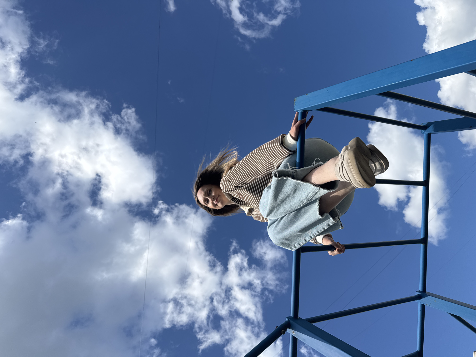 A young woman is pictured sitting on a blue metal jungle gym against a bright blue sky with white clouds. She is wearing a striped long-sleeved shirt and light blue wide-leg jeans. Her left leg is bent, and her right leg is extended. She is looking towards the camera with a slight smile. The jungle gym appears to be in a public outdoor space, possibly a park or playground, in the city of Chaikovsky, Russia.