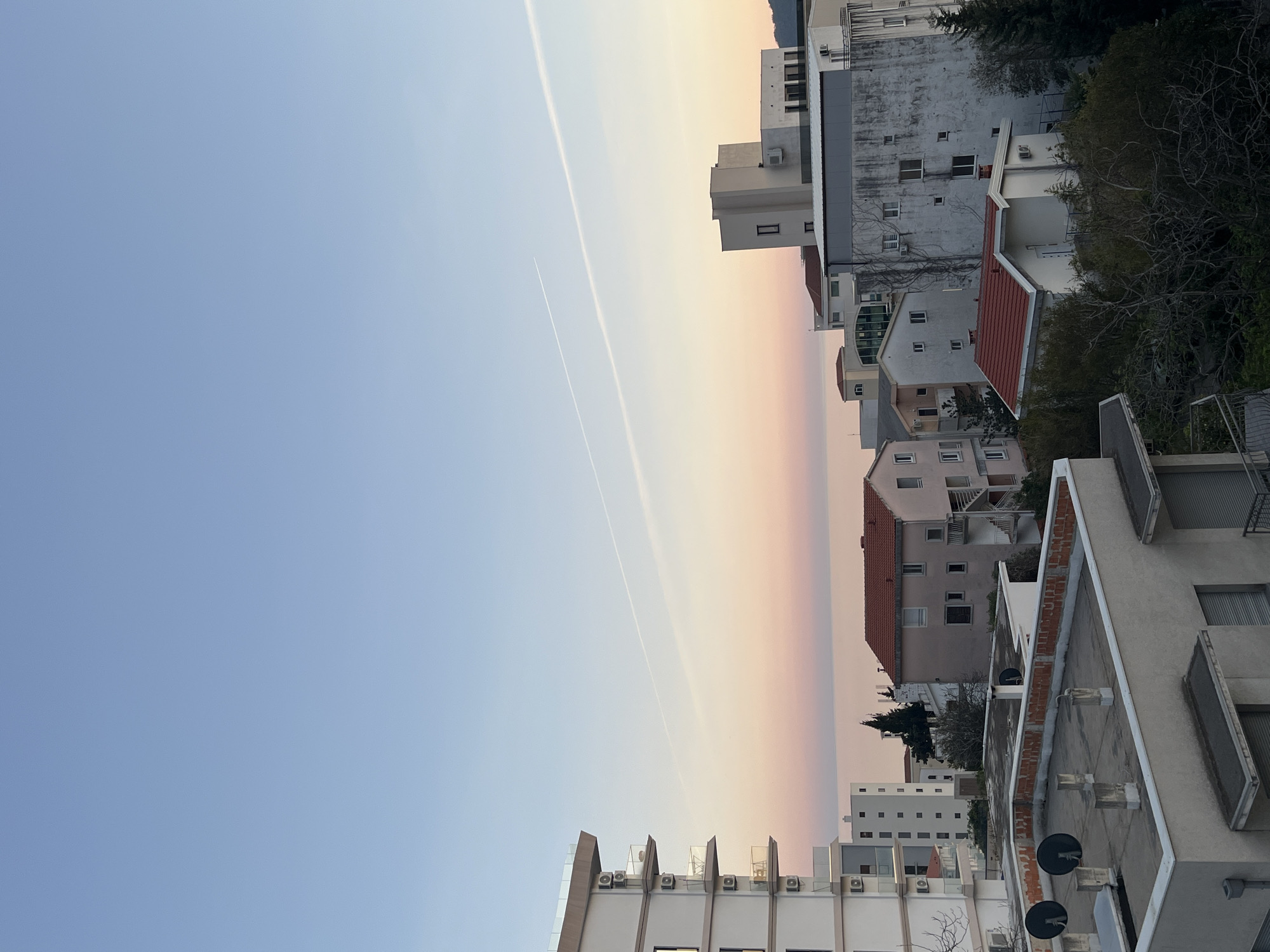 A vertical, eye-level shot shows a view of the sky and buildings in Boreti, Montenegro. The sky transitions from a light blue in the lower left to a pale orange/pink towards the middle. Two contrails are visible diagonally across the sky. Buildings in various shades of white, gray, and peach with some red-tiled roofs fill the right side of the frame. Some greenery is interspersed among the buildings. The buildings are oriented so that the top of the structures are on the right side of the image, which is unusual.