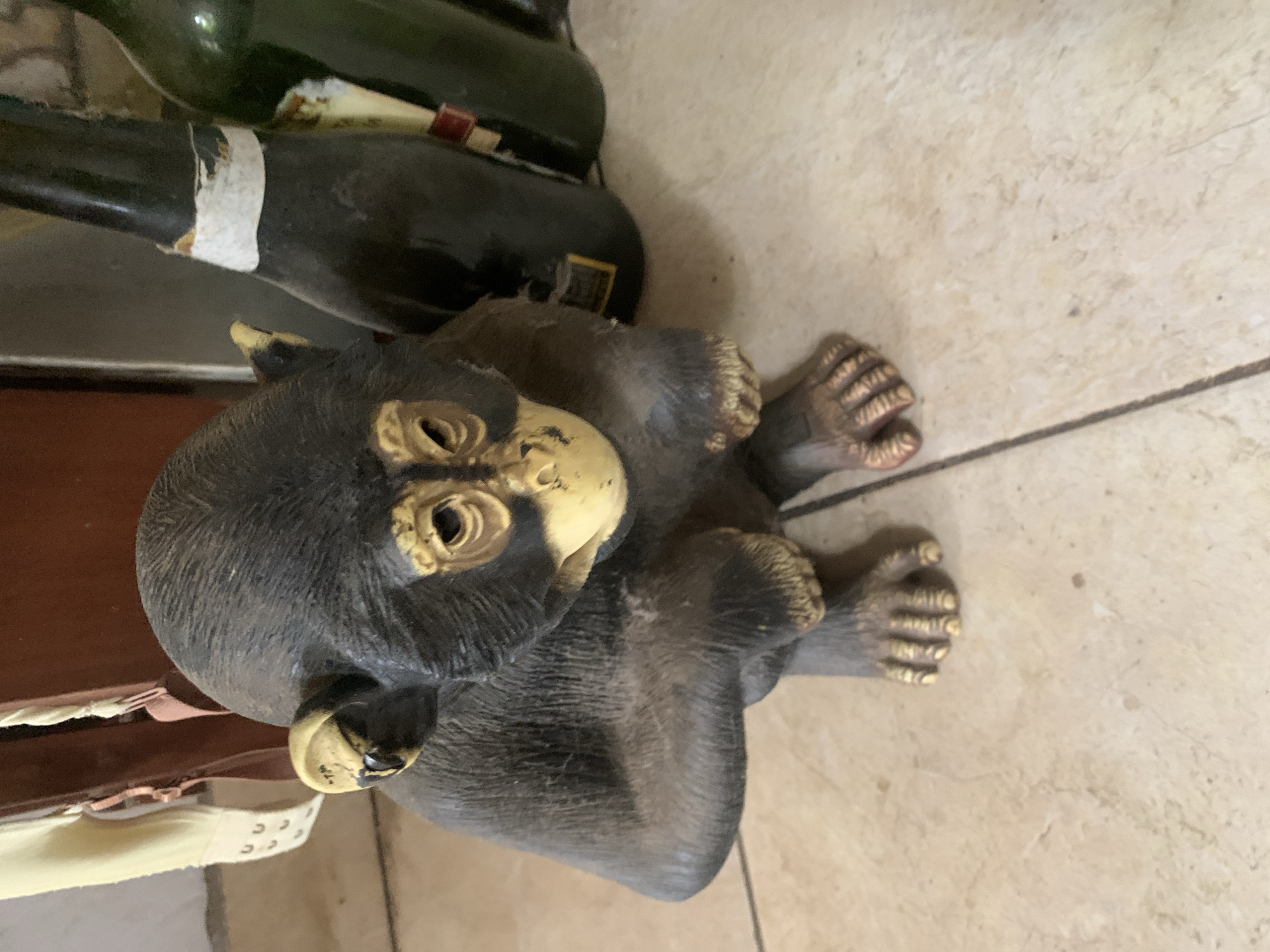 The image displays a small statue of a chimpanzee or monkey in the foreground, situated on a tiled floor. The statue is predominantly dark grey or black, with a lighter, yellowish-gold color on its face, inner ears, hands, and feet. Its eyes appear as dark, empty sockets, and it is depicted in a squatting or sitting pose, looking slightly upward and to the left of the frame.
In the background, two bottles are visible, leaning against a darker surface. One bottle is dark green, while the other is a dark, possibly black, color. The dark bottle has a partially torn, white label wrapped around its neck, and a small yellow label is also visible further down its body. The green bottle also shows hints of a label with some red coloring.
The floor beneath the statue and bottles is made of light-colored, possibly beige or cream-colored, square tiles with darker grout lines. The overall scene suggests an indoor setting. The image itself is rotated 90 degrees clockwise, causing the objects to appear on their sides relative to a standard upright orientation. No specific activity is taking place as the subjects are inanimate objects. No readable text is discernible on the bottle labels or elsewhere in the image. While the location context of Santa Ana, Venezuela is provided, there are no visual cues within the image to specifically identify this location.