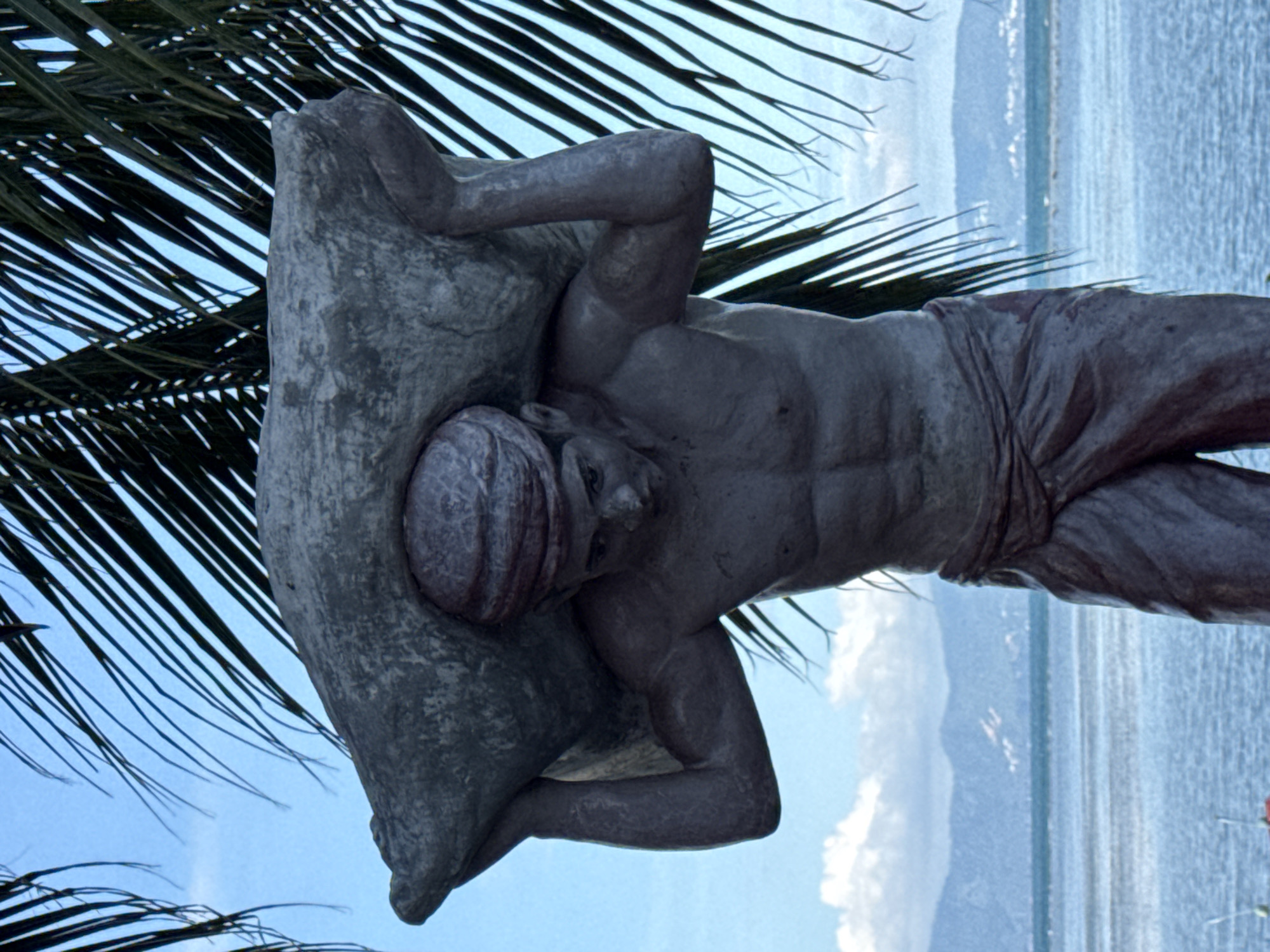 A statue of a man, appearing to be a laborer, is depicted carrying a large sack over his shoulders. The statue is weathered and made of stone. The man is muscular, with a cloth wrapped around his head like a turban and a draped cloth around his waist. He is positioned in front of a body of water, likely the sea, with a distant coastline and mountains visible. Palm fronds frame the top of the image, suggesting a tropical location. The sky is blue with some clouds. The overall impression is of a scene set in a coastal area of Thailand, possibly in the district of Ko Pha Ngan, given the mention of ตำบลเกาะพะงัน.