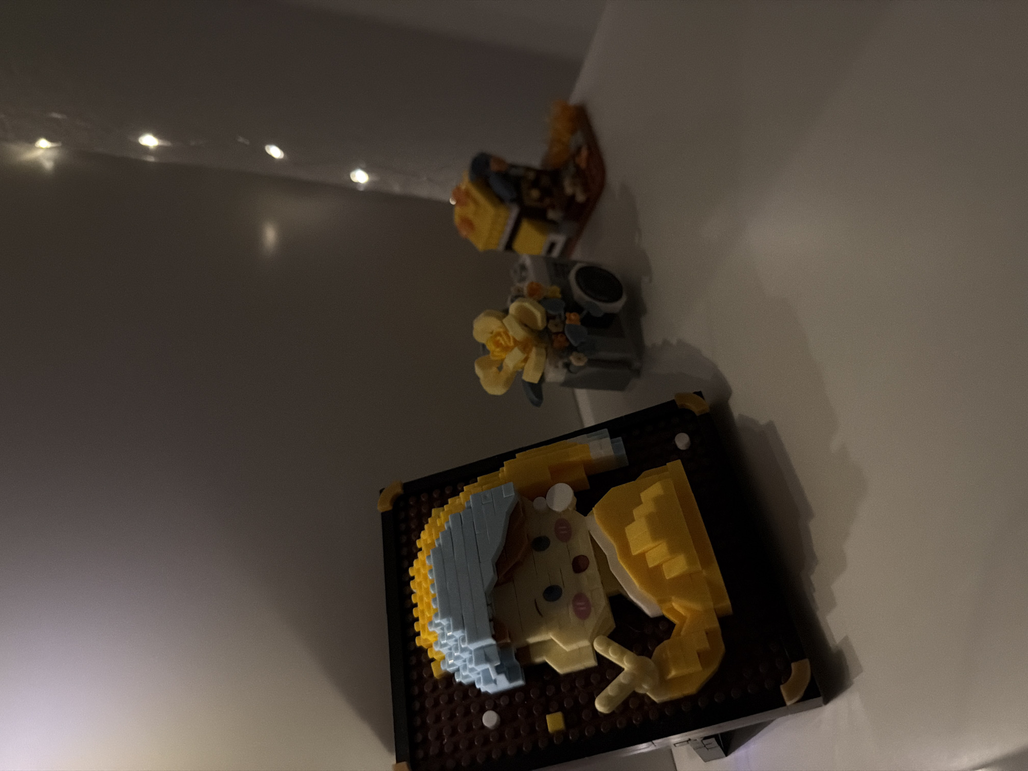 The image depicts a collection of miniature brick models displayed on a white surface. The primary subject is a brick art piece resembling a portrait of a young woman wearing a blue headscarf and a yellow garment, with a prominent pearl earring. To the right of this portrait, there is a miniature brick model of a vintage camera adorned with yellow flowers. Further to the right, a small brick model of a yellow house with a blue roof is visible. The background is dark and features a string of small, blurred lights, suggesting ambient lighting. The scene is shot from a slightly elevated angle, with shadows cast on the surface, indicating a light source from the top left. The overall impression is one of a curated display of hobbyist creations. There is no information in the image to suggest its location in Kyiv, Ukraine.