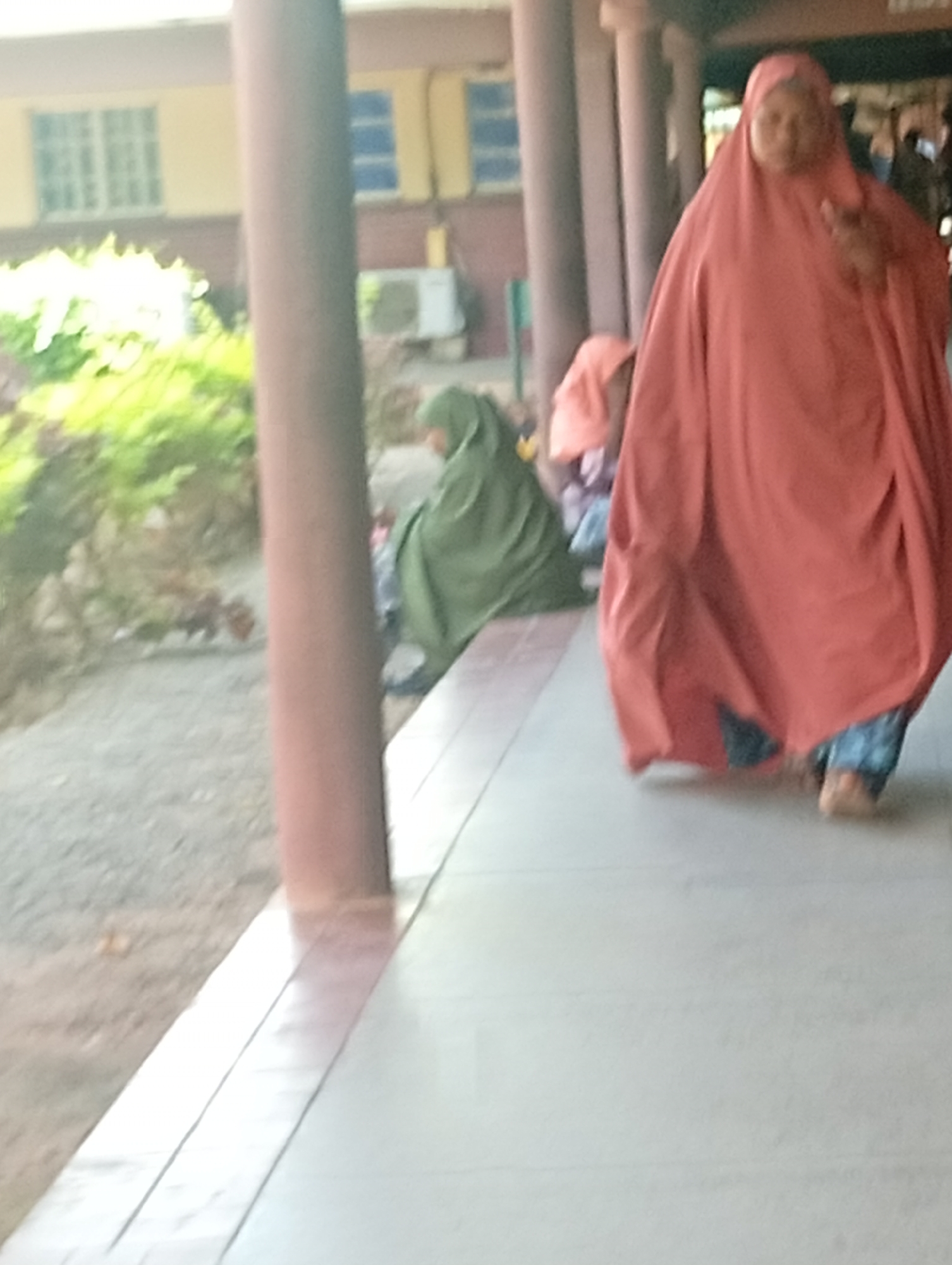 In Gombe, Nigeria, a group of individuals are gathered in an outdoor corridor. The foreground is dominated by a person wearing a voluminous, coral-colored hijab that drapes over their body, obscuring much of their attire. Their feet, clad in blue patterned trousers and appearing bare, are visible beneath the hem of the hijab. To the left, another individual is seated or crouched, wearing a green hijab and a long garment. Behind them, a glimpse of a person in an orange hijab can be seen. The environment features a beige tiled walkway, with a rough, gravelly ground area to the left. A large, reddish-brown pillar stands prominently in the mid-ground. In the background, a building with yellow walls and white-framed windows is visible. An air conditioning unit is mounted on the exterior wall of the building. Green foliage adds a natural element to the scene, suggesting a vegetated area adjacent to the walkway. The overall impression is one of a casual gathering in an outdoor public space.