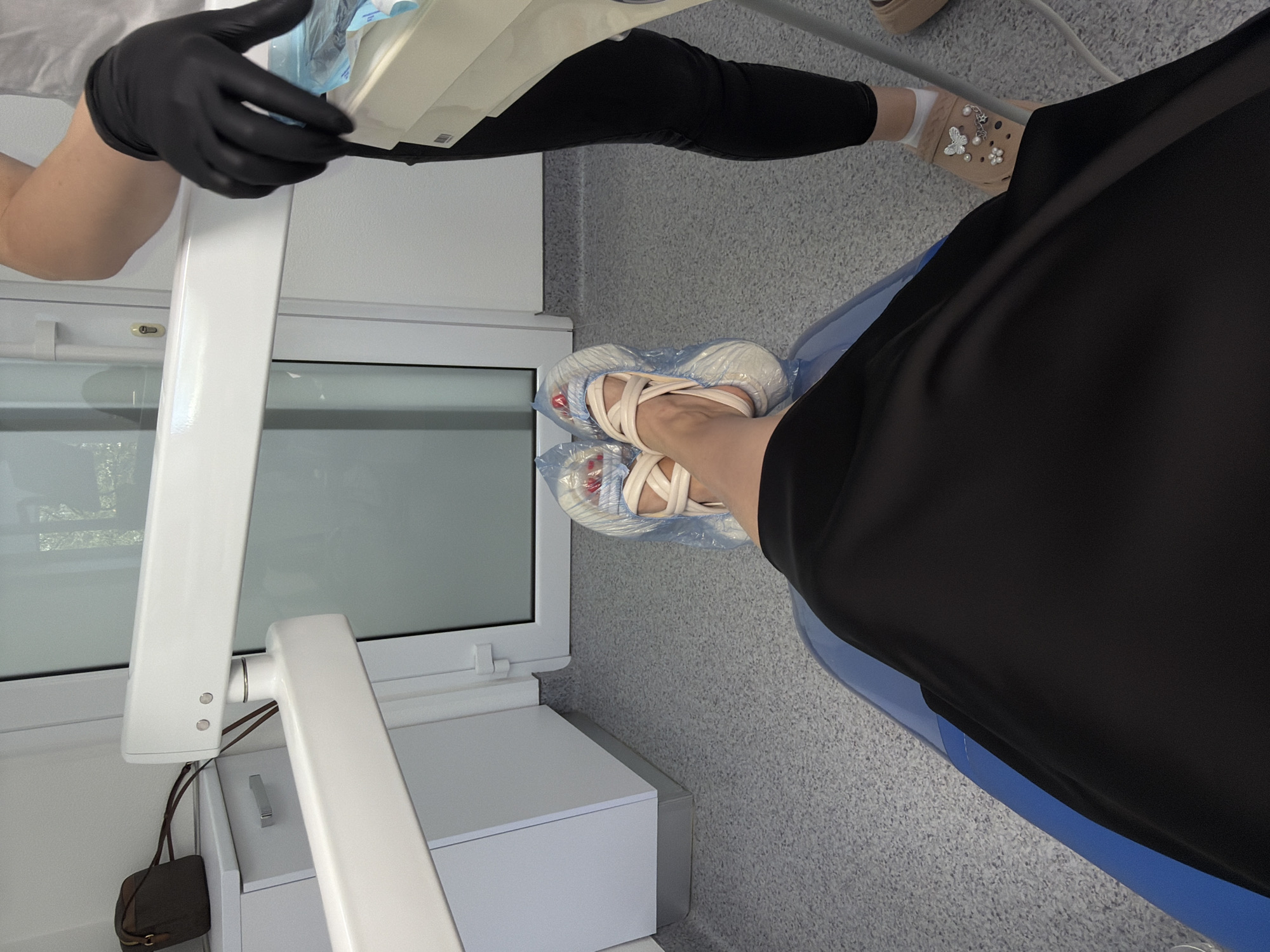The image shows a first-person perspective of a person sitting in a dentist's chair. The person's legs are visible, wearing a black skirt and light-colored sandals with criss-cross straps. The feet are covered in clear plastic shoe covers. To the right of the image, a person wearing black gloves and black pants is standing and holding a package of dental supplies. The environment appears to be a dental clinic, with white dental equipment and light gray flooring visible. A small brown purse is seen on a white cabinet in the background.