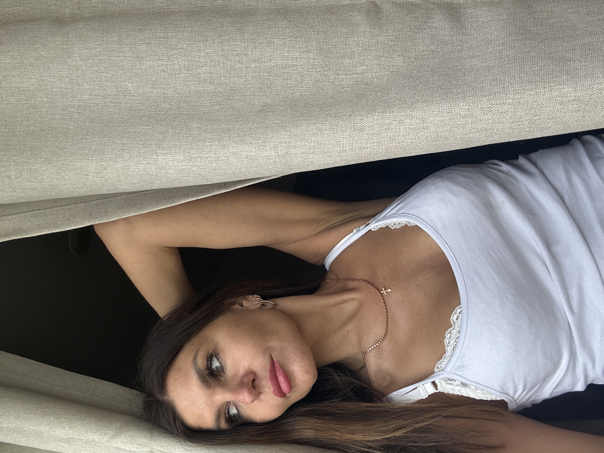 A woman with long brown hair and wearing a white tank top with lace trim is lying down, seemingly between two beige cushions or surfaces. She has fair skin, a necklace with a cross pendant, and is looking to the side with a slight smile. Her arm is raised above her head, and the background is a dark, undefined space. There is no evidence to determine the specific location.
