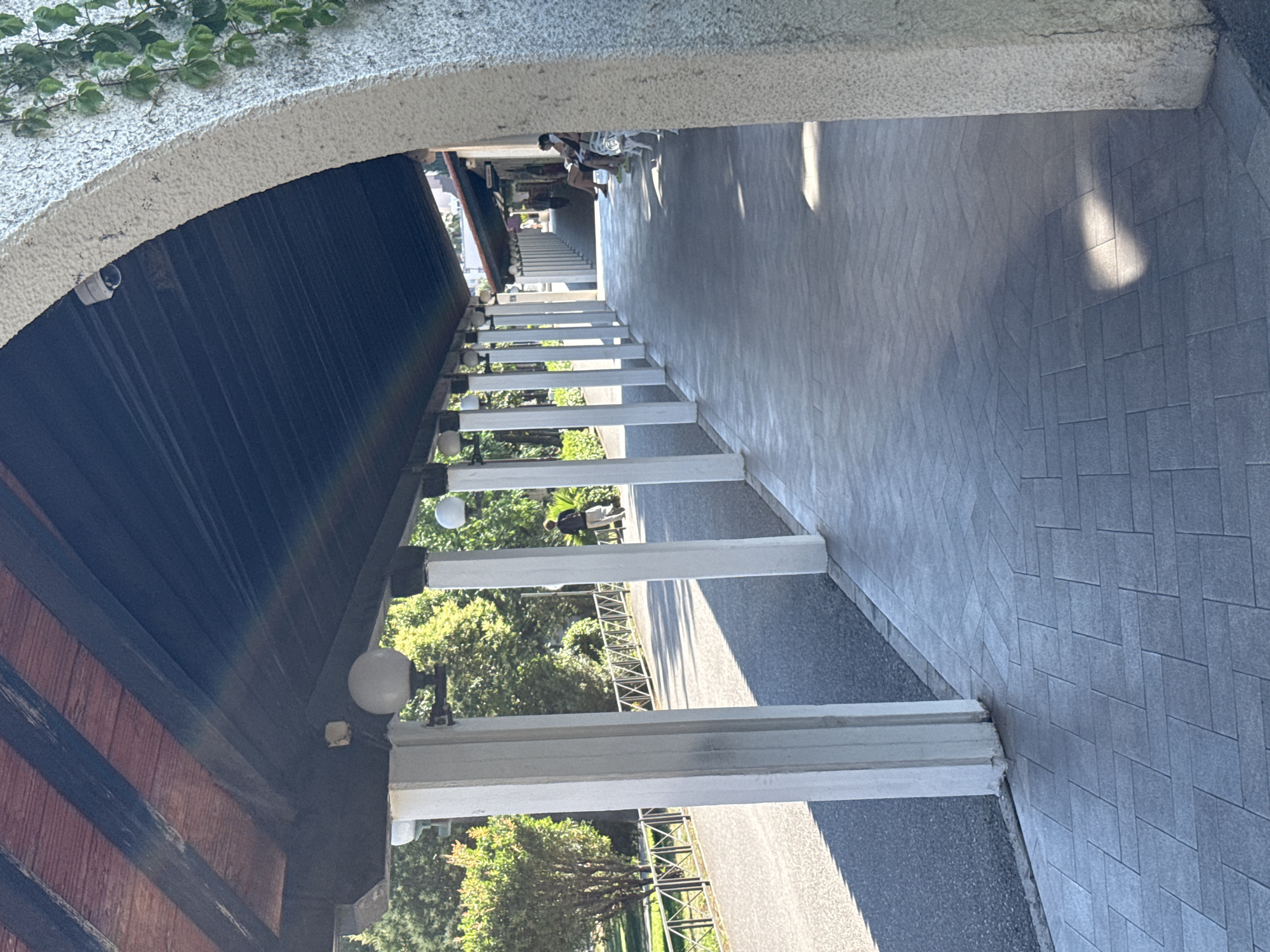 The image depicts an outdoor scene in Budva, Montenegro, with a focus on an architectural structure resembling a covered walkway or an archway leading to an outdoor seating area. The perspective is from within the covered area, looking outwards.
In the foreground, on the left, is the underside of a textured, light-colored curved roof or ceiling, with green ivy growing on its outer edge. Below this, there's a dark, slatted wooden structure, possibly part of the ceiling or wall, with some red-stained wood visible. Several white, rectangular pillars or supports run vertically down the walkway, leading the eye towards the exterior. Between these pillars, glimpses of greenery from trees and foliage can be seen. On the right side of the walkway, the ground is paved with rectangular gray tiles arranged in a patterned fashion. Some sunlight creates distinct light and shadow patterns on this tiled surface, suggesting it is daytime.
Further into the scene, beyond the pillars, there's an outdoor seating area with white chairs and tables. A few people are seated there, appearing relaxed and engaged in conversation or simply enjoying the environment. One person is standing near the seating area, facing away from the camera. The overall atmosphere seems tranquil and leisurely, possibly indicating a cafe or restaurant setting. In the background, more trees and a glimpse of buildings suggest an urban or semi-urban environment. The time of day appears to be daytime, likely late morning or afternoon, given the strong sunlight and shadows. There is no visible text in the image.