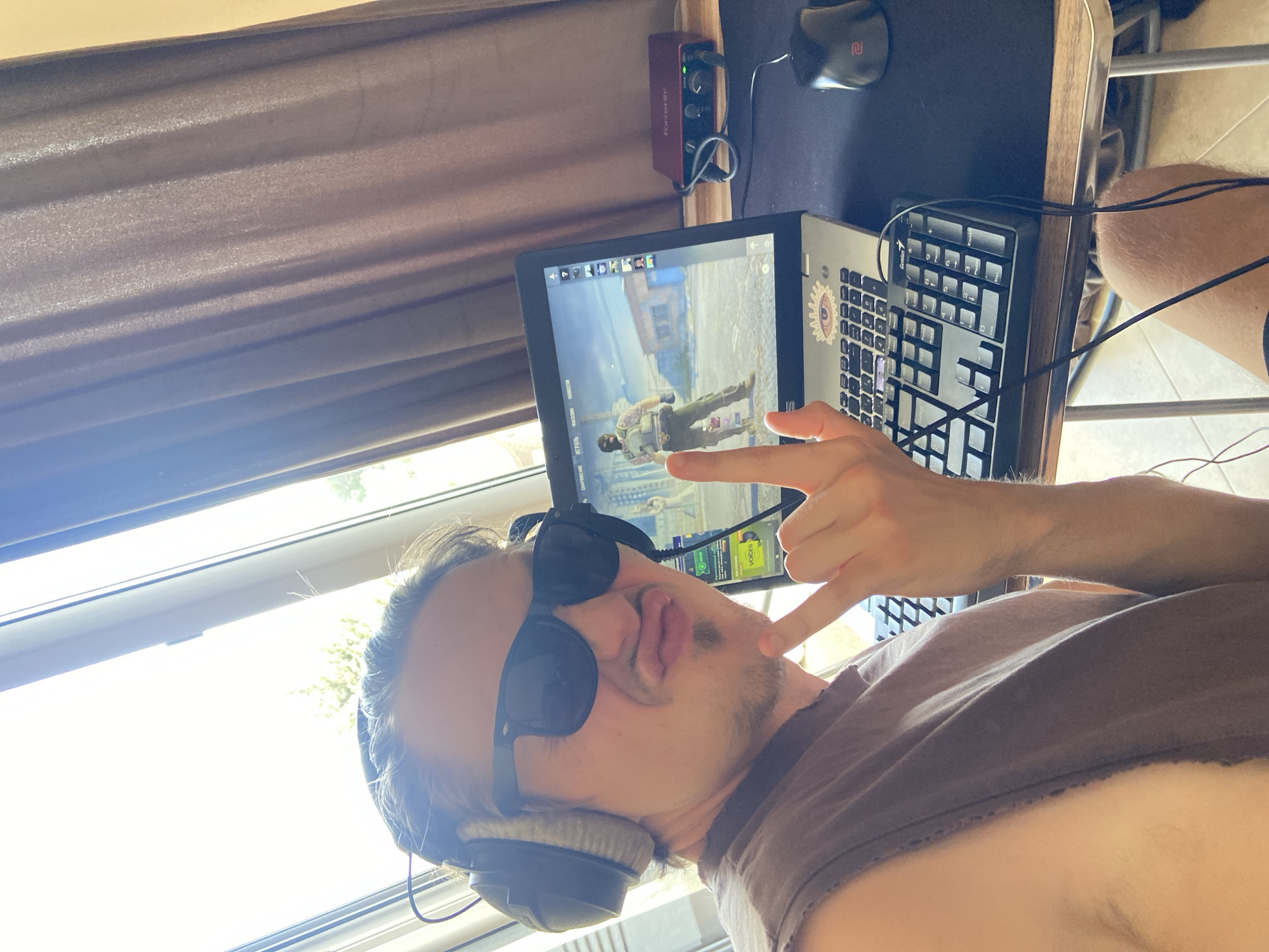 A person wearing sunglasses and headphones is taking a selfie while sitting at a desk. A laptop displays a video game with a first-person perspective, a character appearing in the center of the screen. A black keyboard and a black mouse are on the desk. A red audio interface is attached to the side of the desk. The scene appears to be inside a building with a window and a curtain in the background. The location is not identifiable as Šušanj, Montenegro based on the image content.