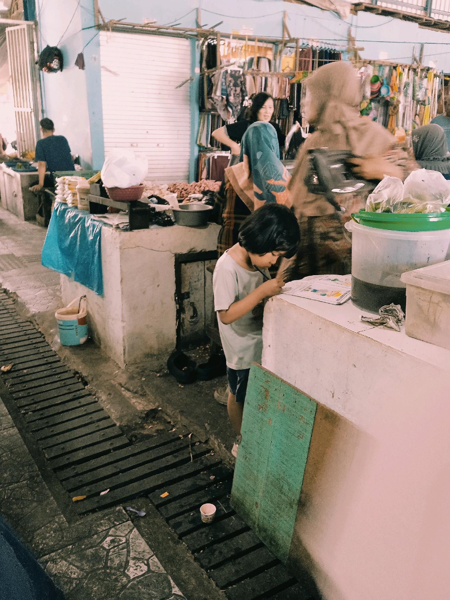 This image depicts a street market scene in Banjarmasin, Indonesia. In the foreground, a young child with dark hair, wearing a light blue t-shirt and dark shorts, is leaning over a counter, engrossed in looking at a booklet or piece of paper. Next to the child, on the counter, are a large clear plastic tub filled with dark liquid and a smaller white container. A green bucket is visible on the ground next to the counter, and further left, a person wearing a dark blue t-shirt is seen from behind, standing near a stall with goods displayed. The background reveals more market stalls with clothing and other items hanging. The environment is somewhat cluttered and appears to be an outdoor or semi-outdoor market. The ground is a mix of paving stones and a slatted metal grate.