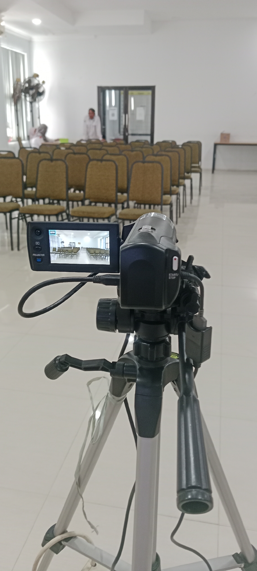 The image shows a video camera mounted on a tripod in a room with rows of empty chairs. The camera's LCD screen displays a view of the room, showing more chairs and a table in the background. In the far background, two people are visible, one standing and one seated. The room appears to be a meeting or conference space. The overall lighting is bright, suggesting the use of artificial lights or ample natural light. The floor is tiled and appears to be clean. The camera and tripod are the primary subjects in the foreground. The scene is located indoors, likely in Tuban, Indonesia, given the context of the user's query.